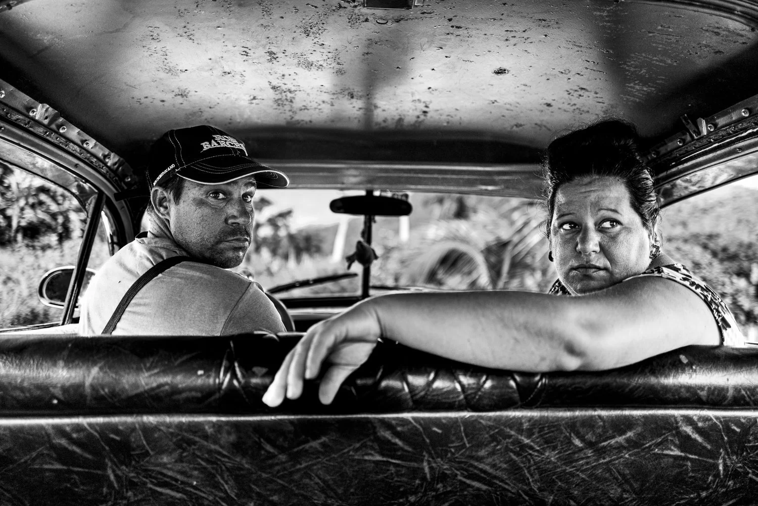 Black-and-white portrait of two people looking back from inside a vintage car in Cuba.