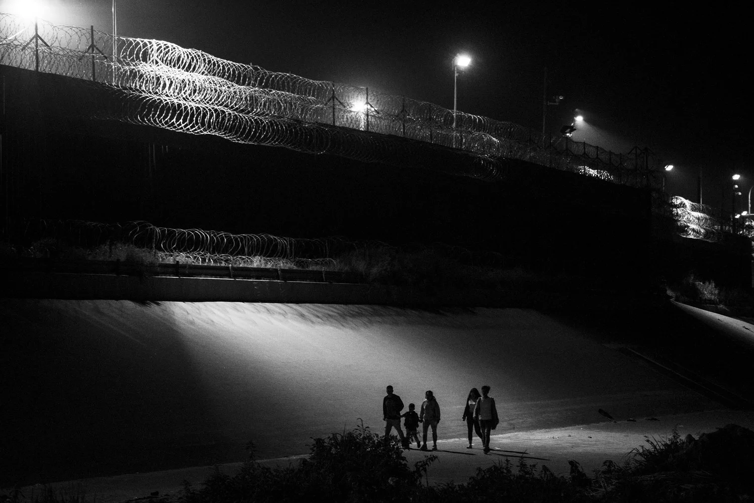 Black-and-white photograph of a family helping one another along the riverbank at the Juárez–El Paso border at night.