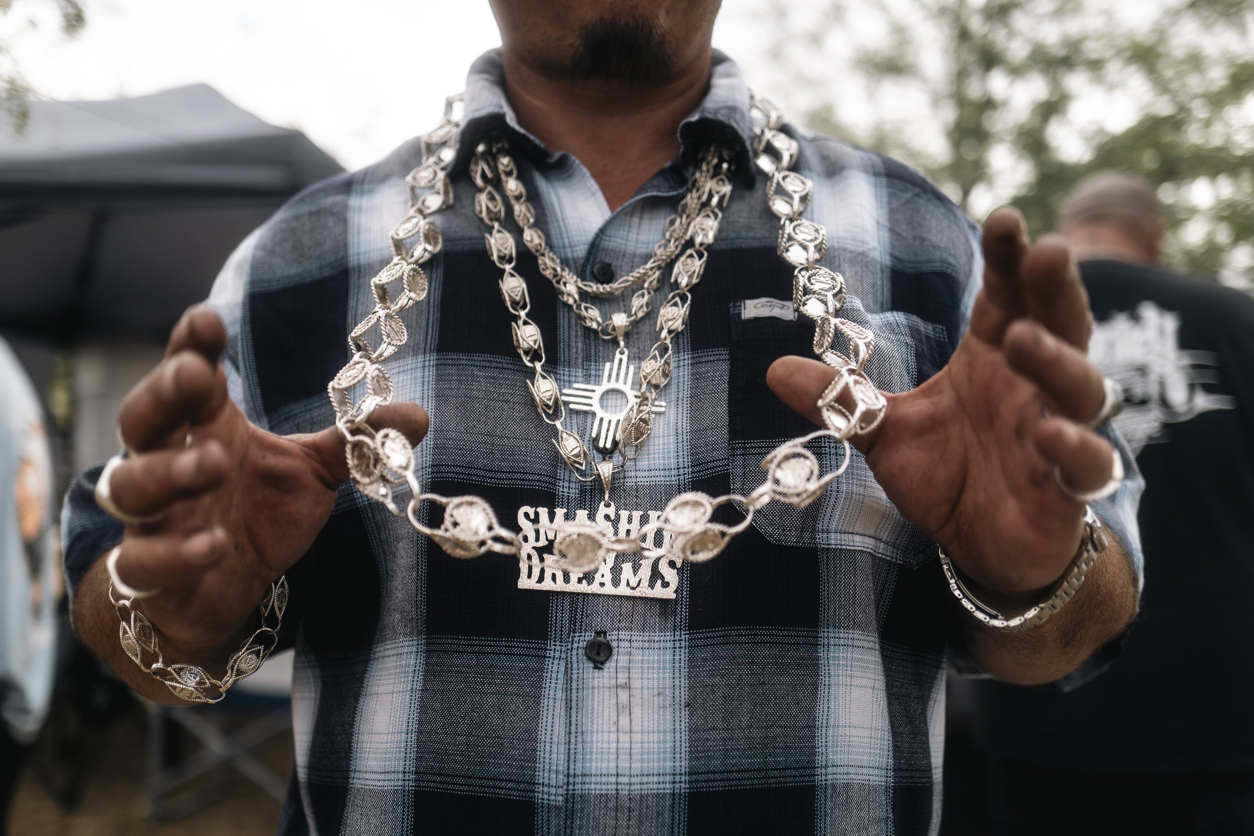 A man wearing a blue plaid shirt showing off multiple silver chains and jewelry, including necklaces, rings, and a bracelet, with a 'S m o k e Hous e D e a l e r s' pendant, at an outdoor gathering.