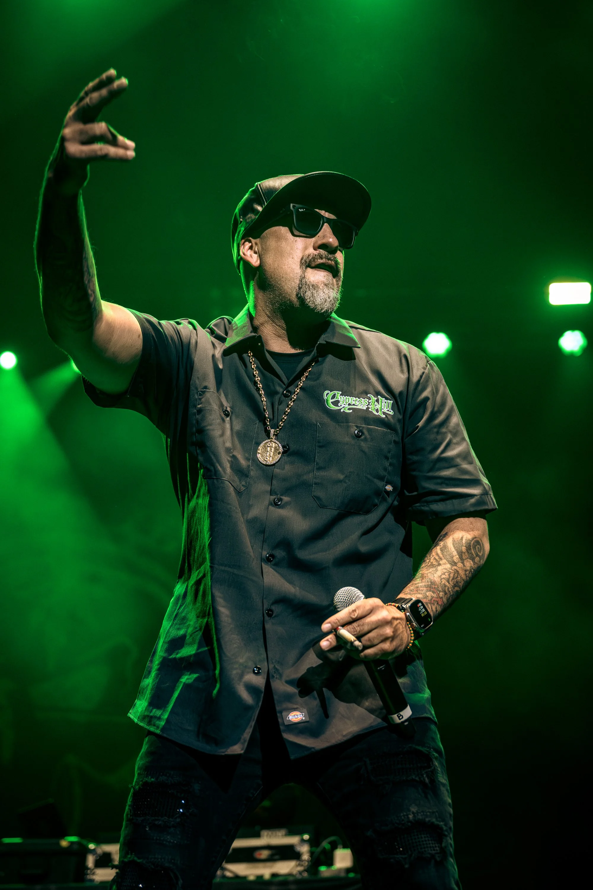 A man with tattoos, wearing a black cap, sunglasses, a dark short-sleeve shirt with embroidery, and a gold chain, holding a microphone on stage with green lighting in the background.