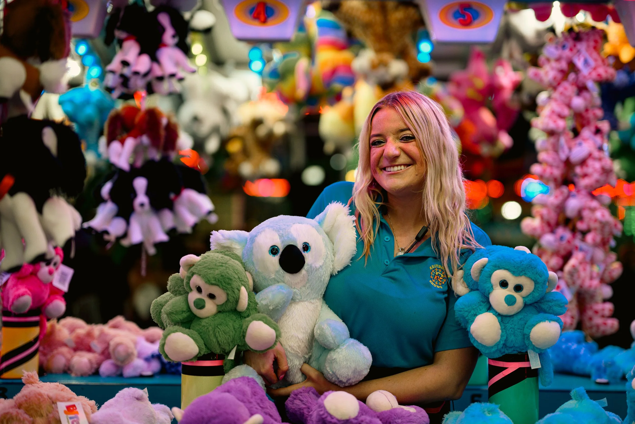 A young woman smiling and holding plush toys at a carnival game booth with colorful stuffed animals displayed on shelves behind her.