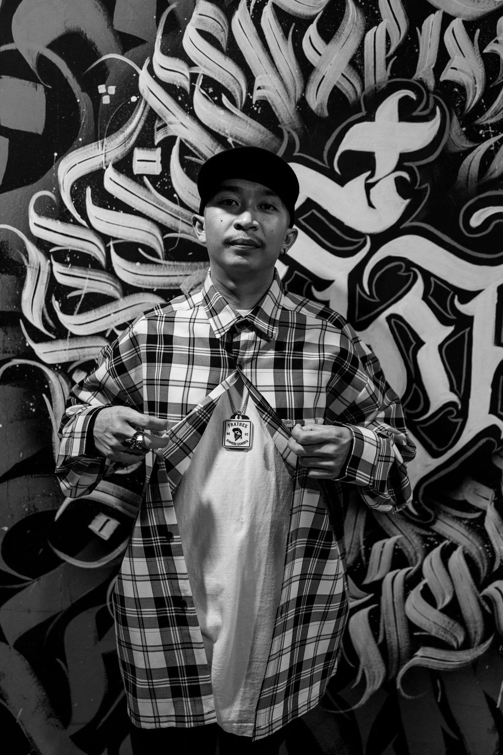 A young man standing in front of a graffiti wall, pulling open his plaid jacket to reveal a shirt underneath with a large pendant necklace. He is wearing a cap and has rings on his fingers.