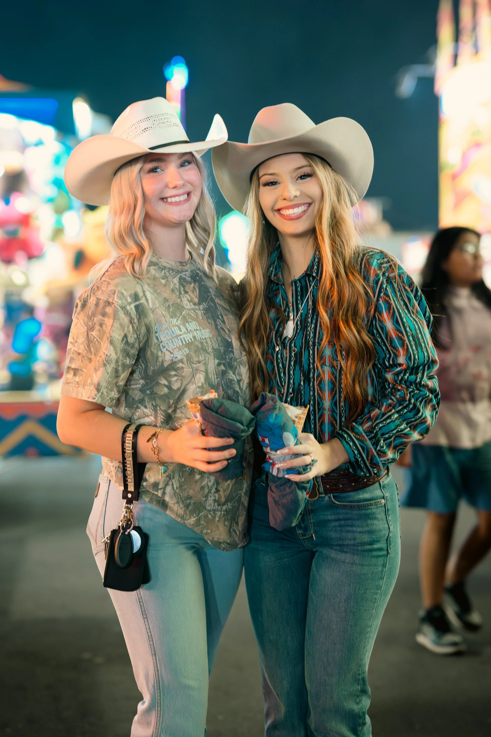 Two young women at a fair wearing cowboy hats, standing close together and smiling. One has blonde hair and wears a camouflage t-shirt, the other has long wavy hair and wears a patterned shirt. They hold snacks and stand in front of colorful carnival
