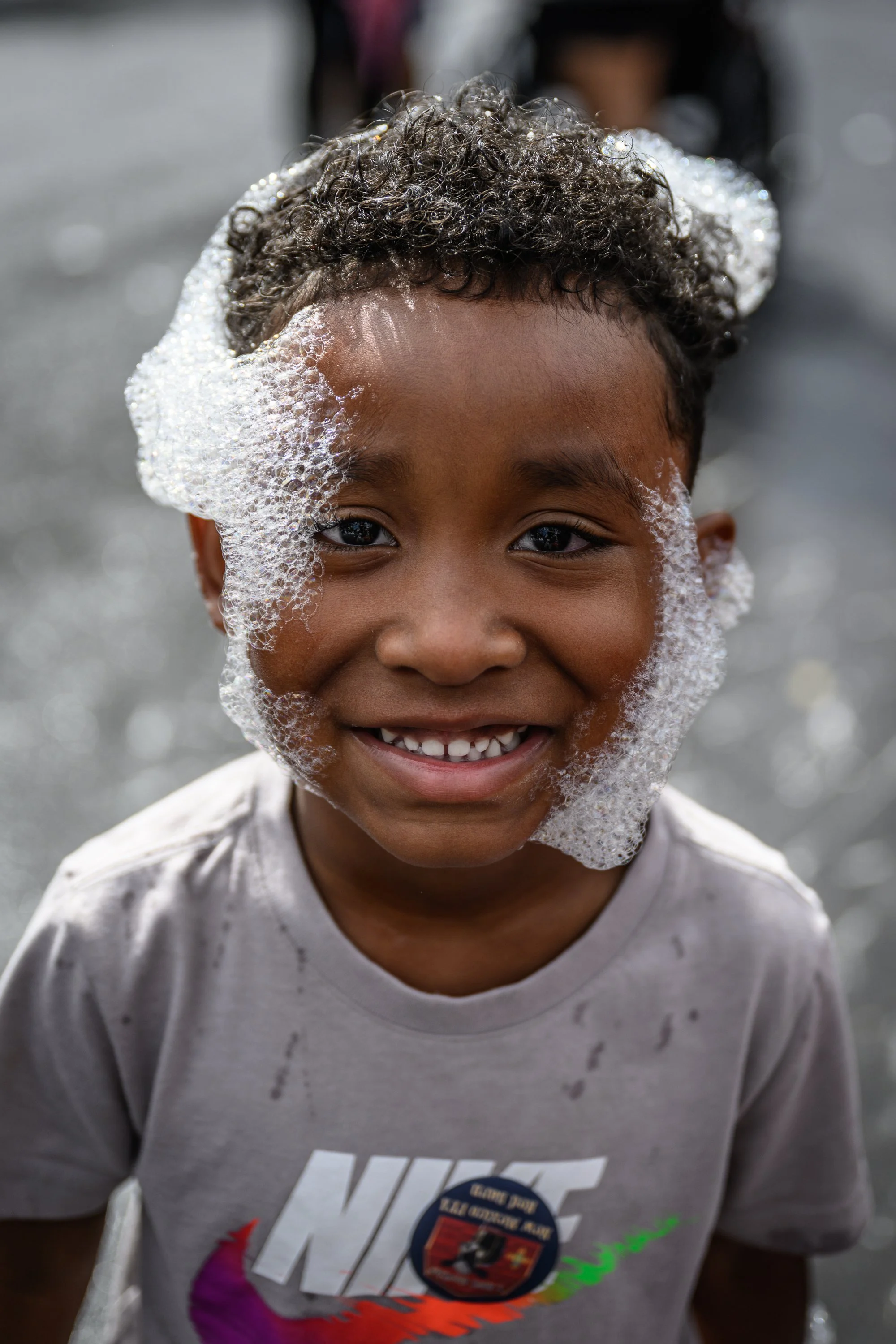A smiling young boy with soap on his face, wearing a gray Nike t-shirt with a badge and rain stains, standing outdoors on a wet surface.