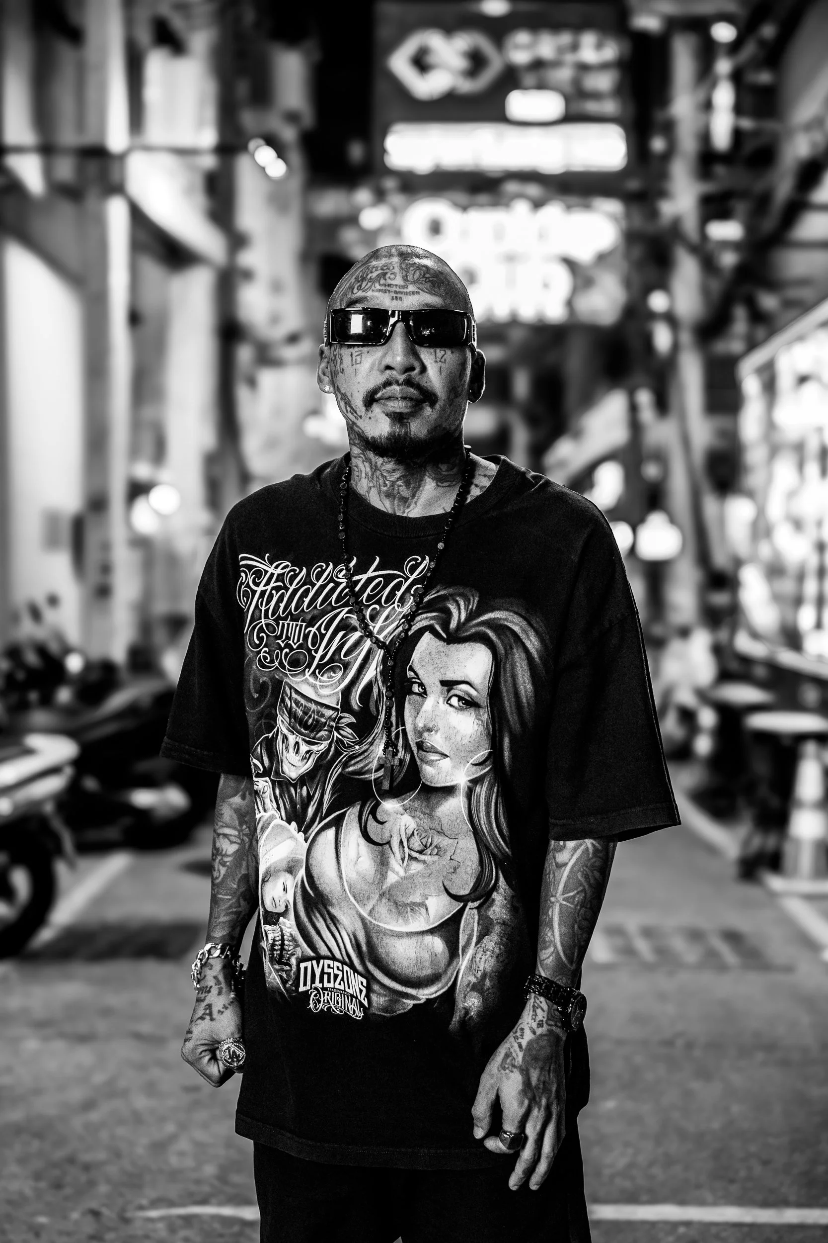 A man with numerous tattoos and sunglasses standing in an urban setting, wearing a graphic T-shirt with a woman’s face and other artwork, in black and white photography.