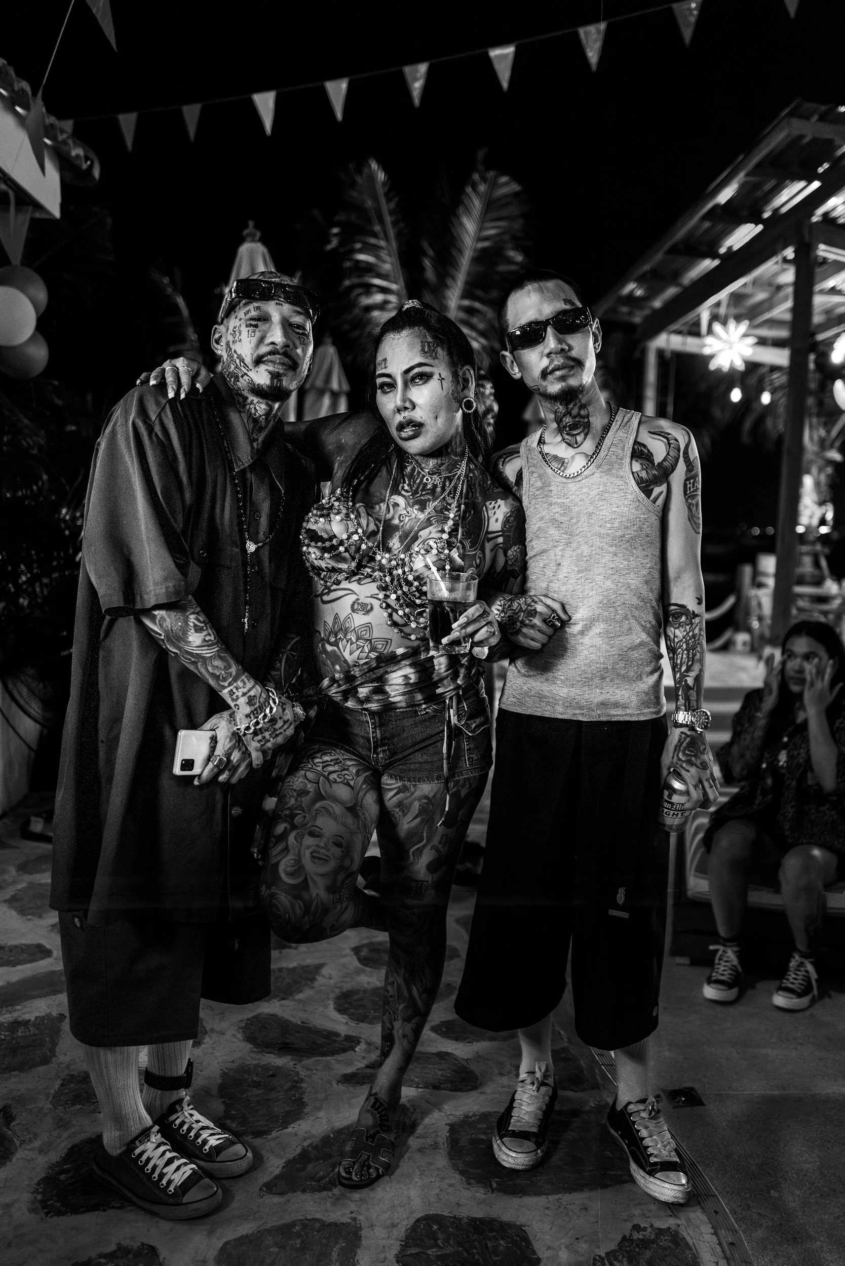 Three tattooed people at a nighttime party, standing close together. The woman in the center has numerous tattoos and is holding a drink. The two men on either side are also covered in tattoos, one wearing sunglasses and a tank top, the other in a da
