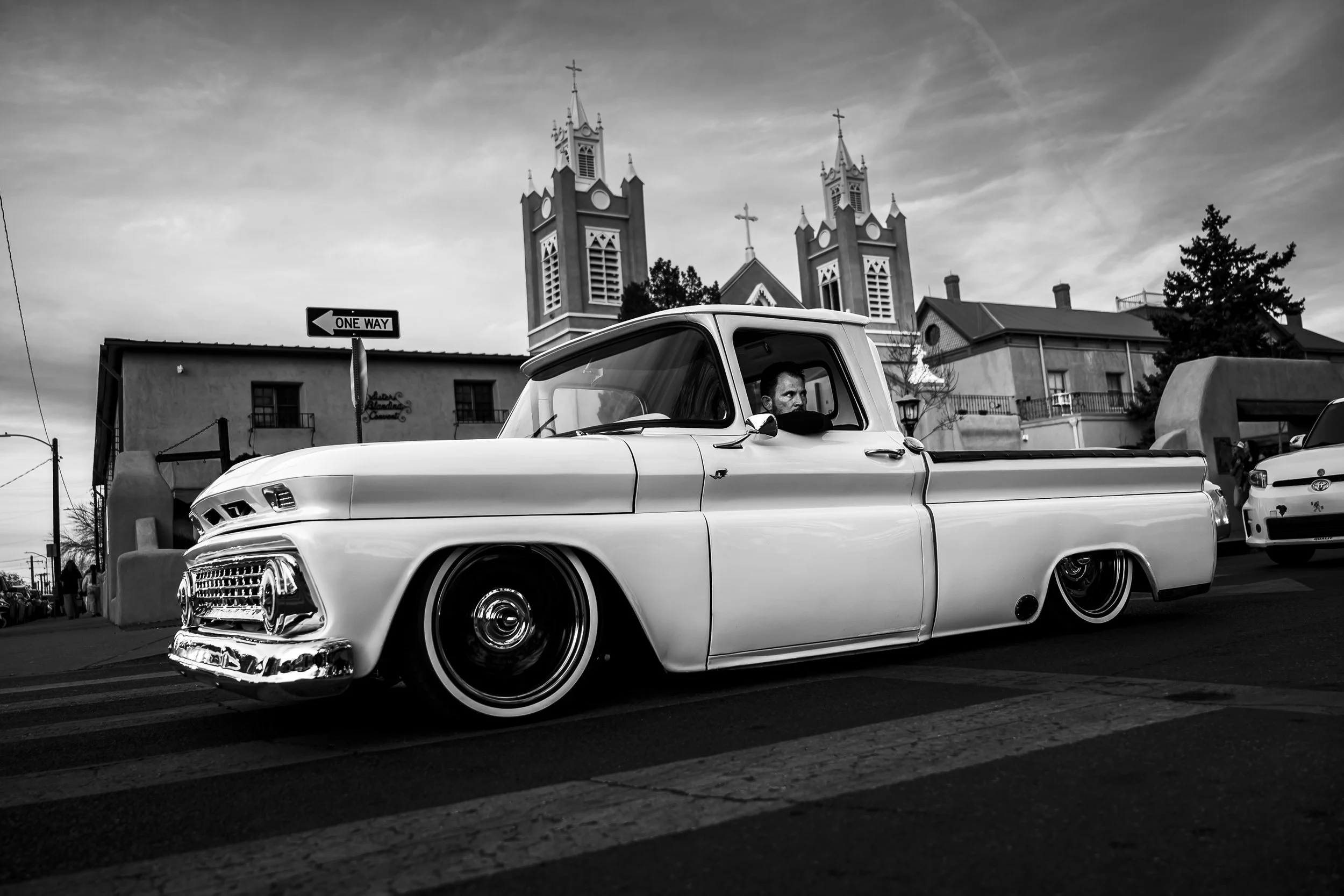 Black and white photo of a vintage lowered pickup truck with a person inside, crossing a street with a church and buildings in the background.