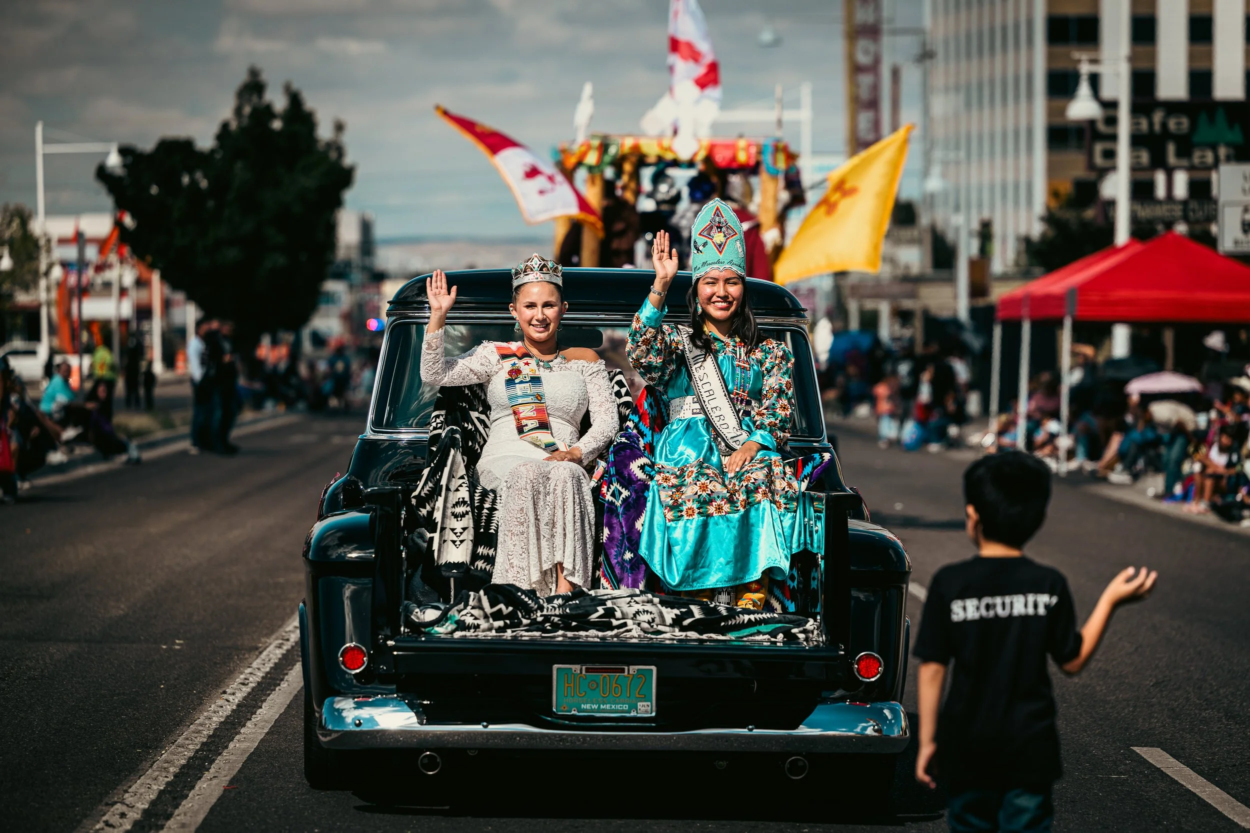 Two women dressed in traditional and festive attire riding in the back of a black vintage truck during a parade, with an audience and flags in the background on a city street.