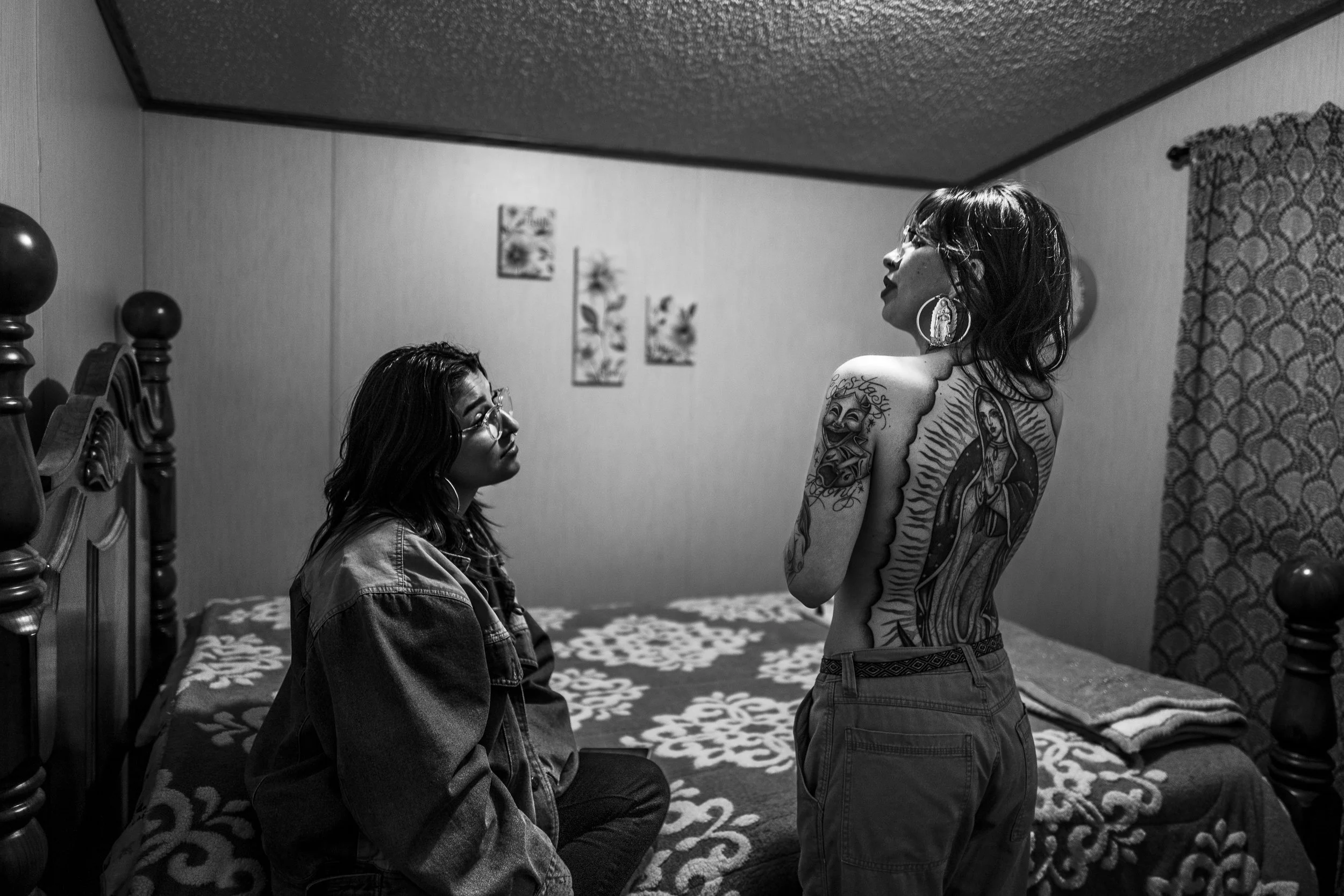 Two women in a bedroom, one sitting on the bed wearing glasses and a denim jacket, the other standing topless with tattoos on her left arm and back, and wearing earrings and glasses. The room has a bed with floral bedding, framed artwork on the wall,