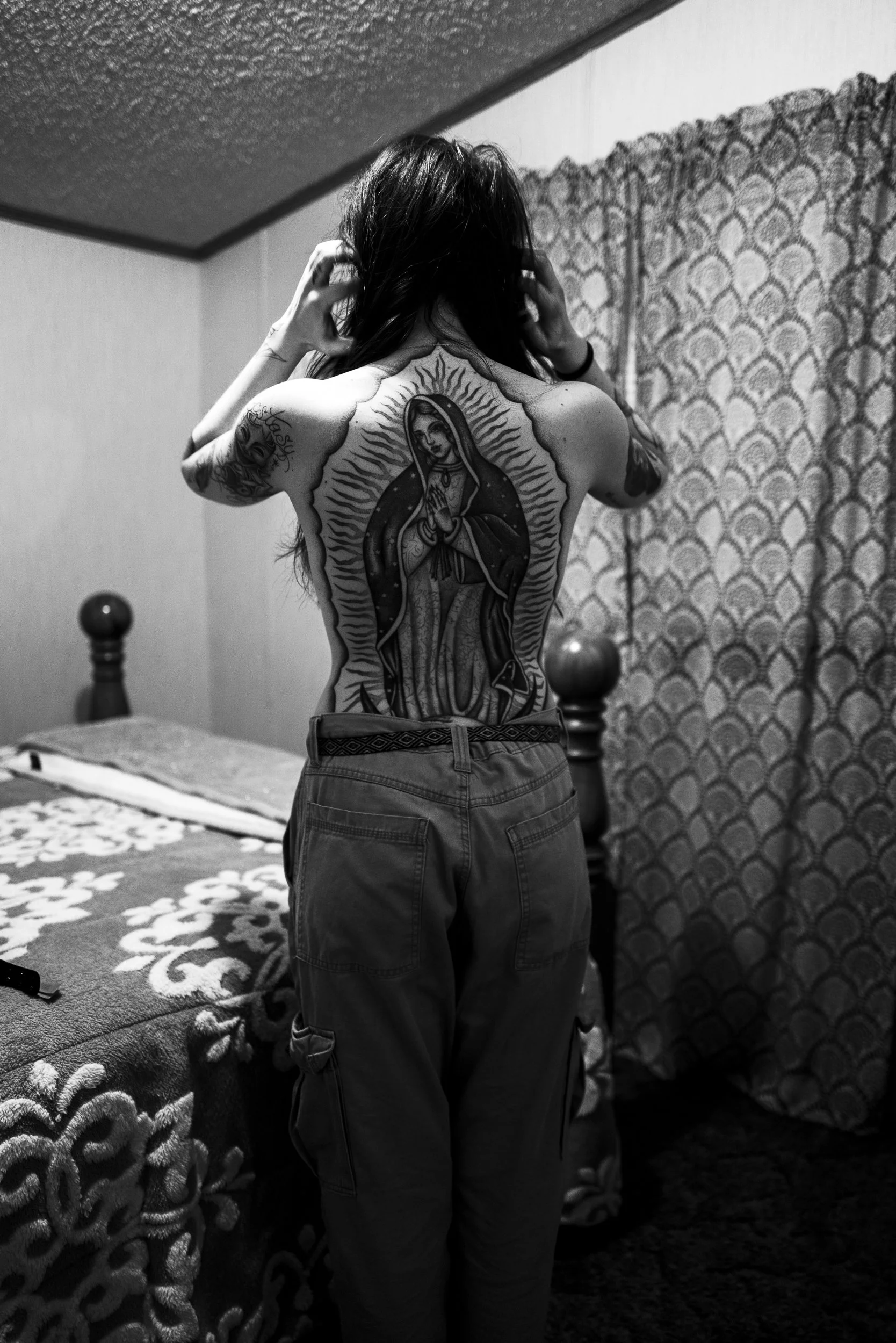 A woman standing in a bedroom, showing her large tattoo of the Virgin of Guadalupe on her back. She has dark hair and is wearing loose pants.