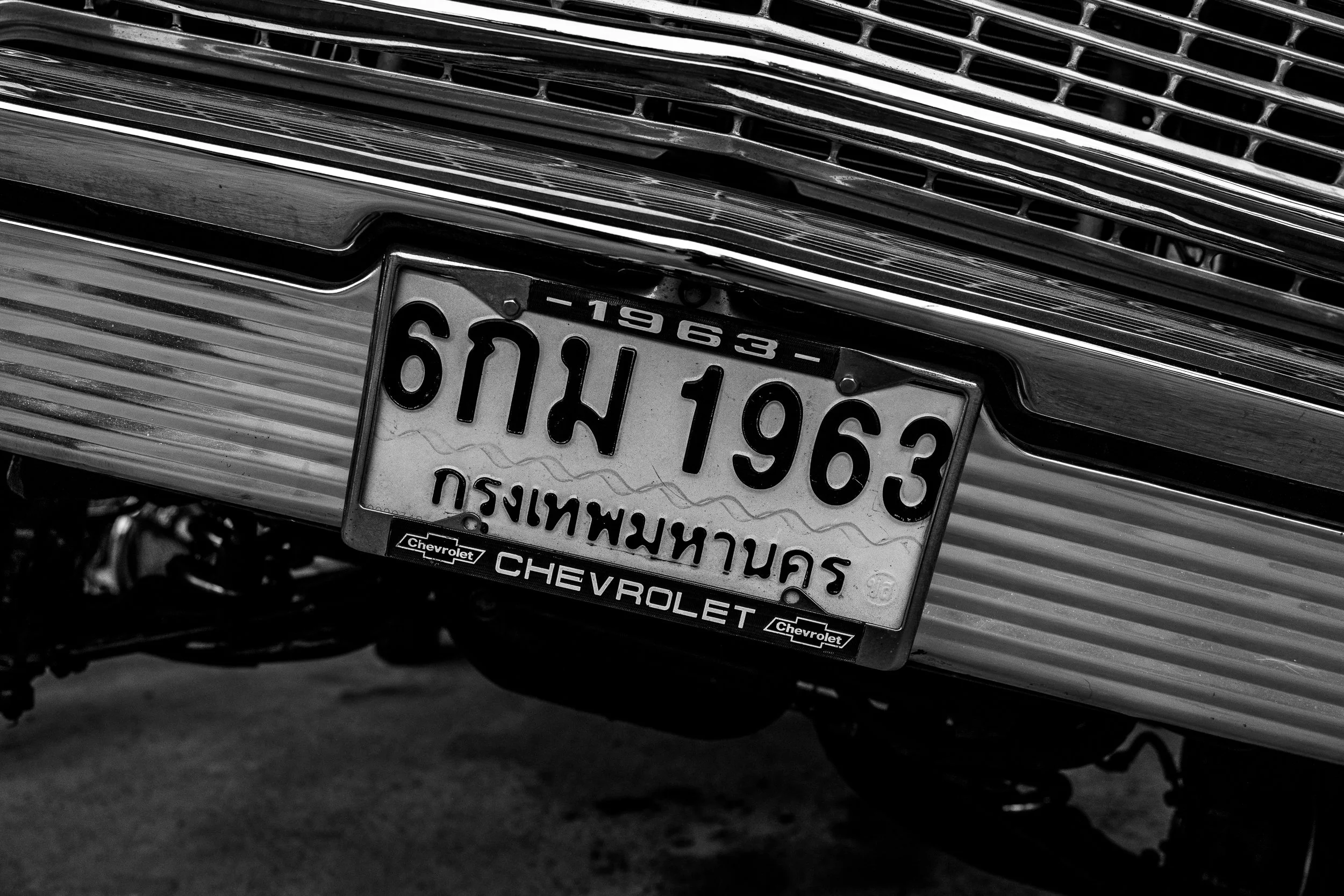 Close-up of a vintage car front with Thai license plate, Chevrolet emblem, and chrome grille in black and white.