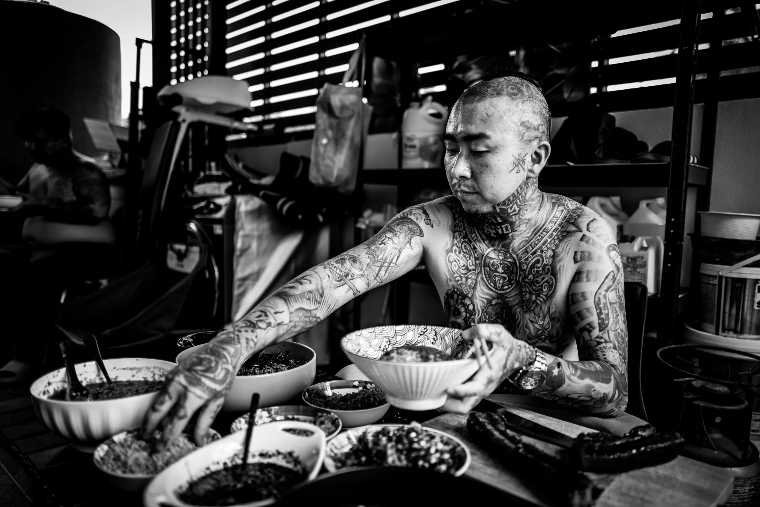A tattooed man with shaved head serving food from bowls at a table in a room with wooden slatted walls.