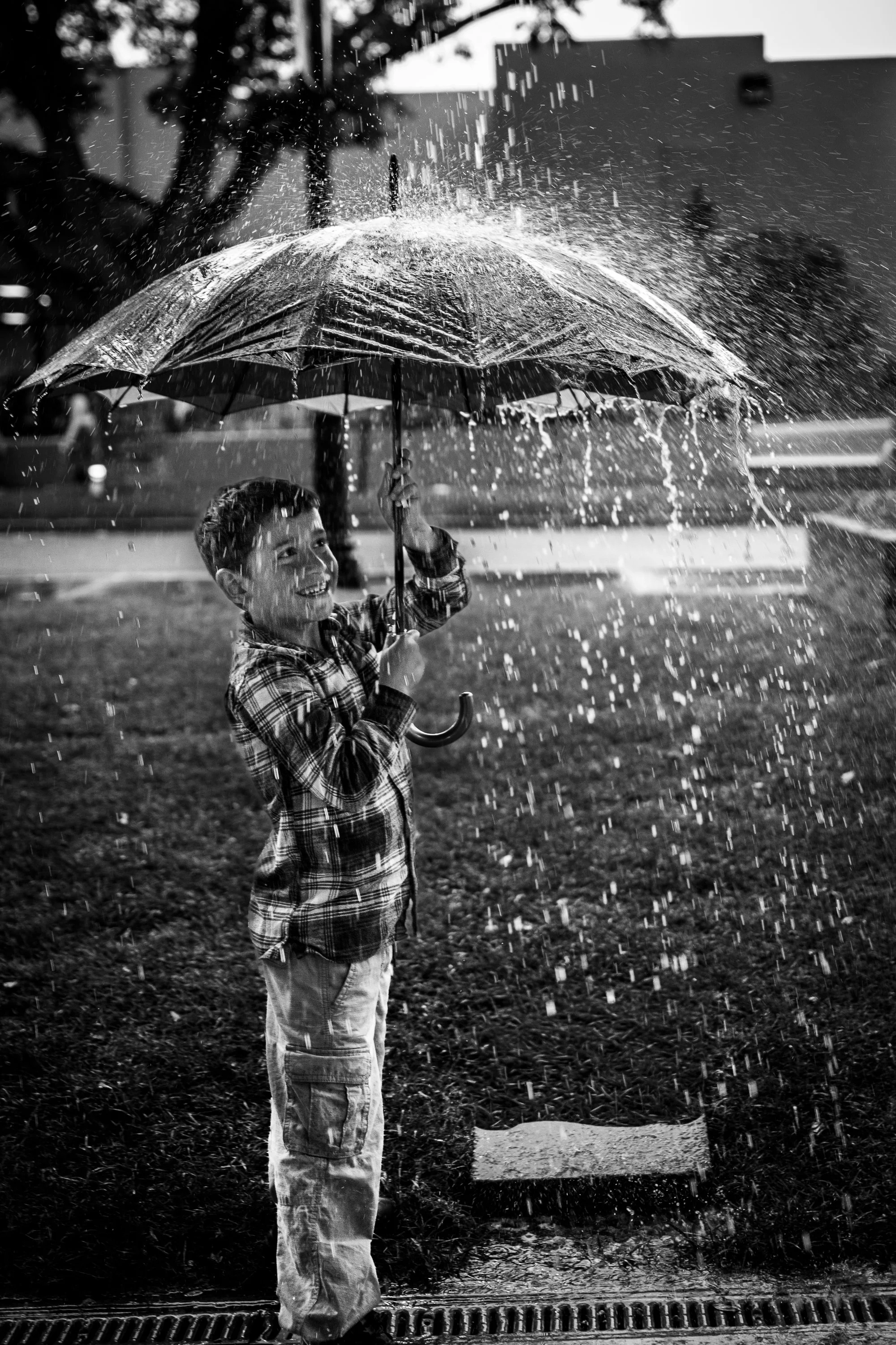A young boy holding an umbrella in the rain, smiling and enjoying the wet weather on a city street.