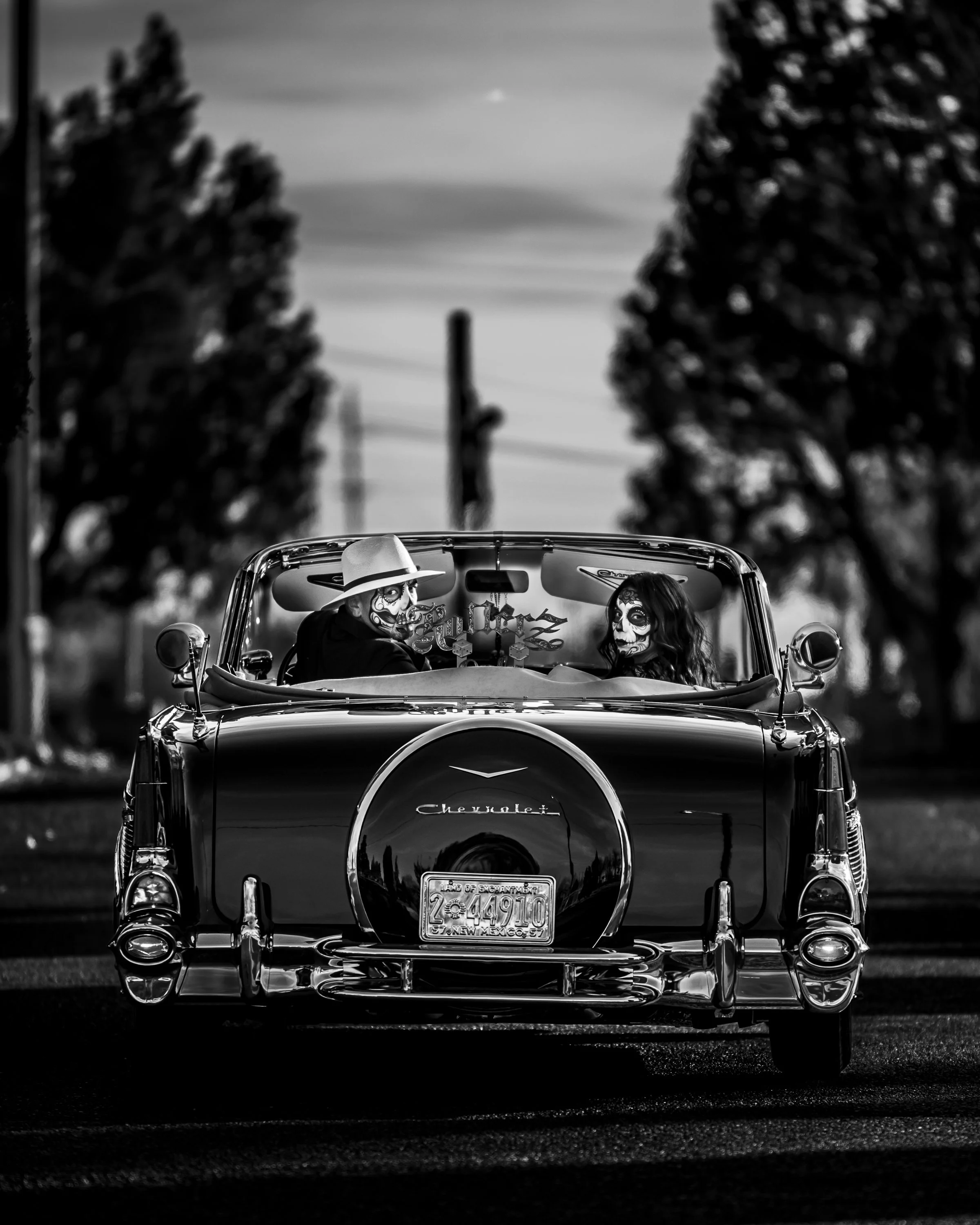 A black and white photo of a vintage Chevrolet convertible car with two people inside wearing day of the dead style face paint and hats, driving down a street with trees and utility poles in the background.