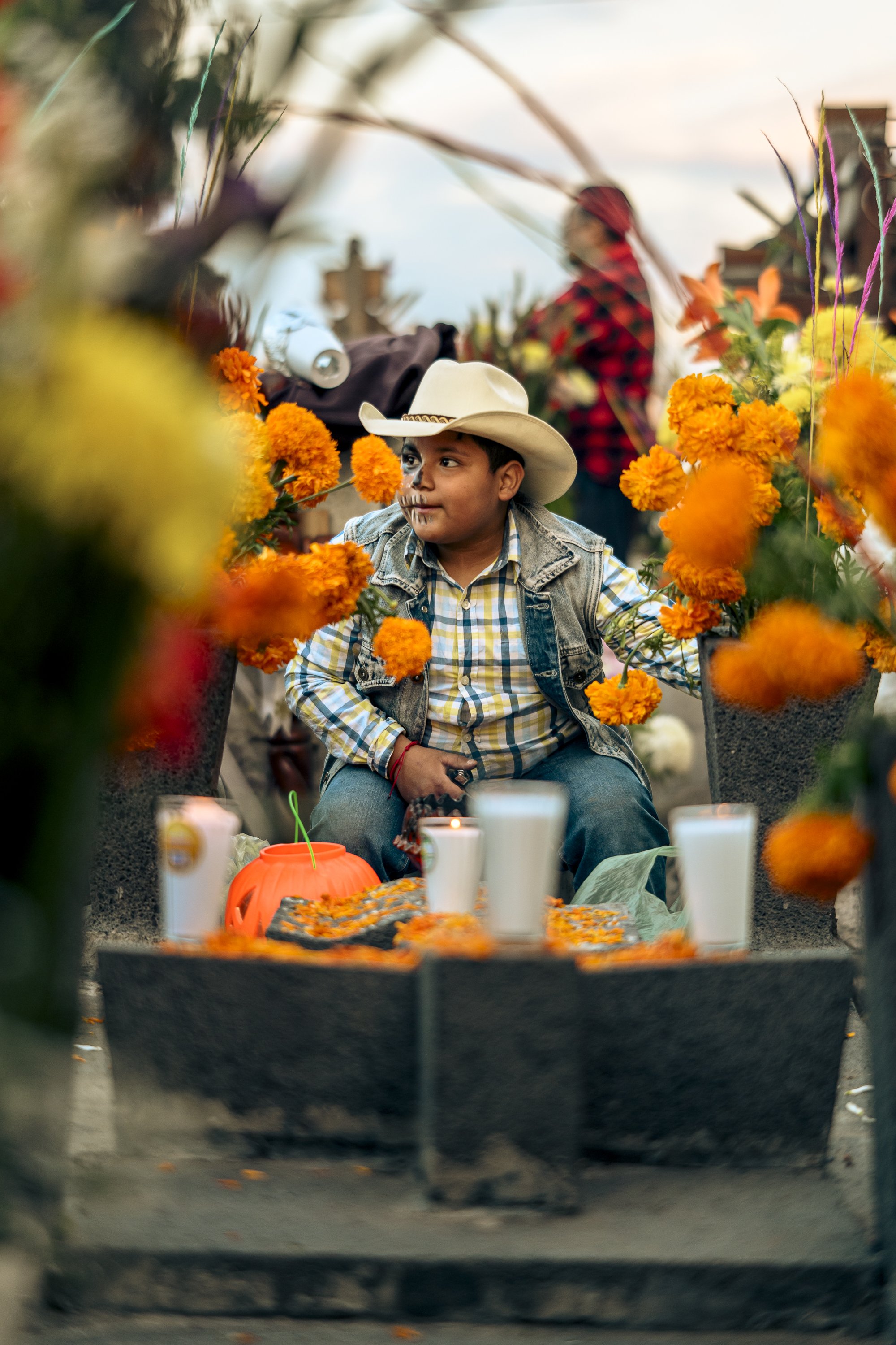 A young boy dressed in a cowboy hat, plaid shirt, and denim vest sitting among orange marigold flowers at a celebration, with candles and a pumpkin in front of him and other people in the background.