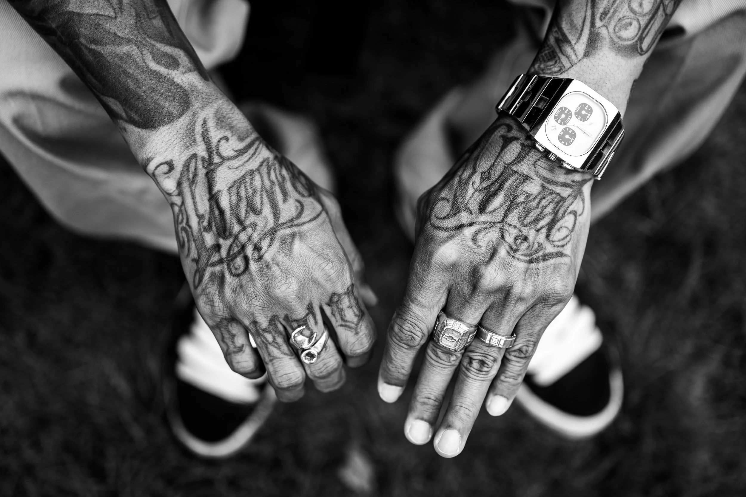 Black and white photo of a person's tattooed hands with rings and watch, taken from above, showing details of tattoos and accessories.