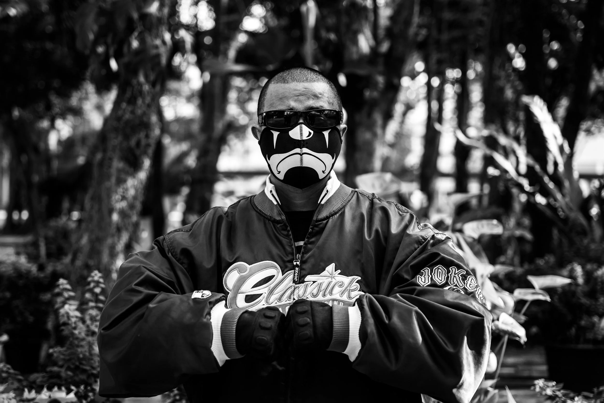 A person wearing a Batman mask, sunglasses, a jacket with the word "Classic" on it, and gloves, standing outdoors with trees and plants in the background.