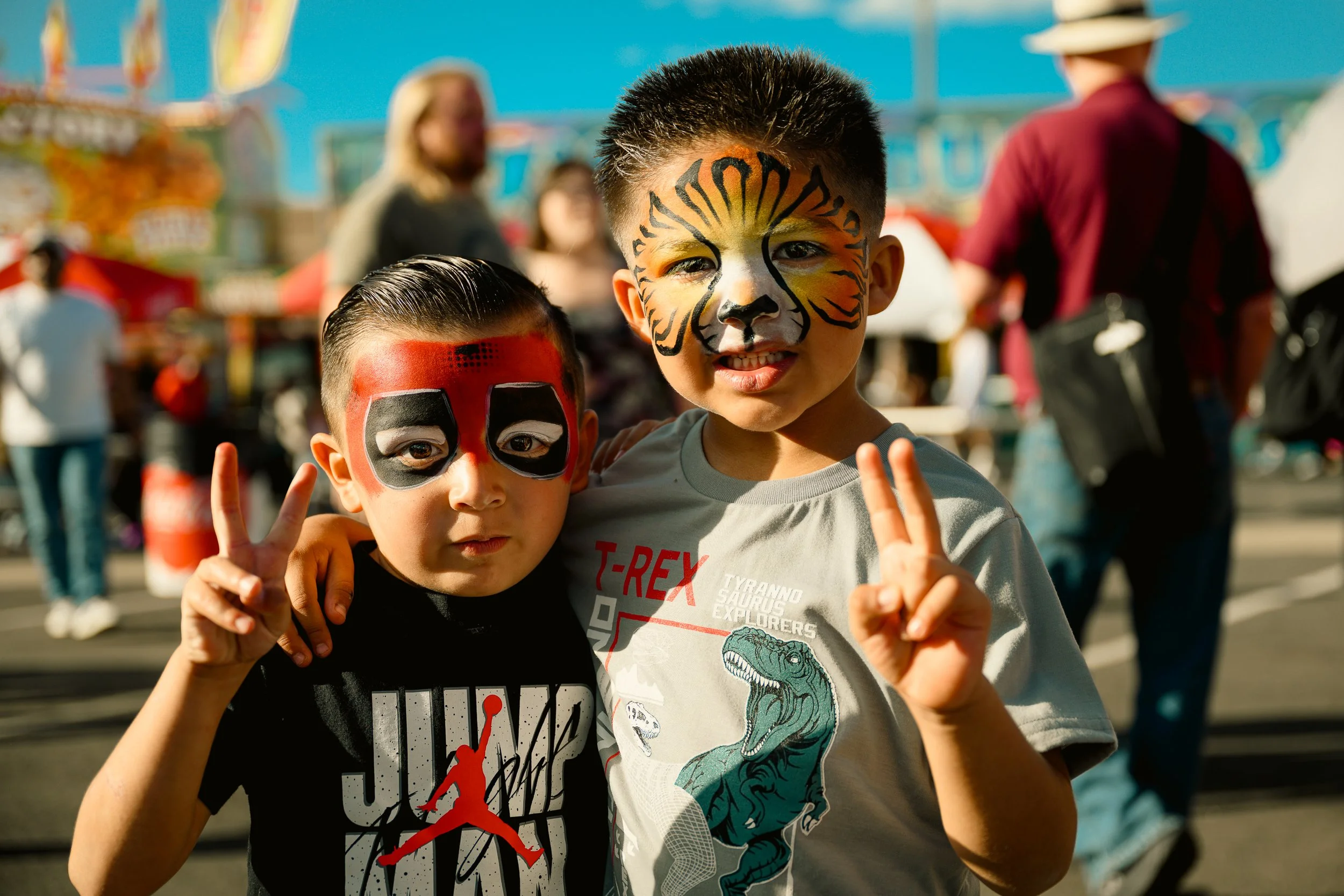 Two young boys with face paint, making peace signs at an outdoor festival or fair, with colorful tents and people in the background.