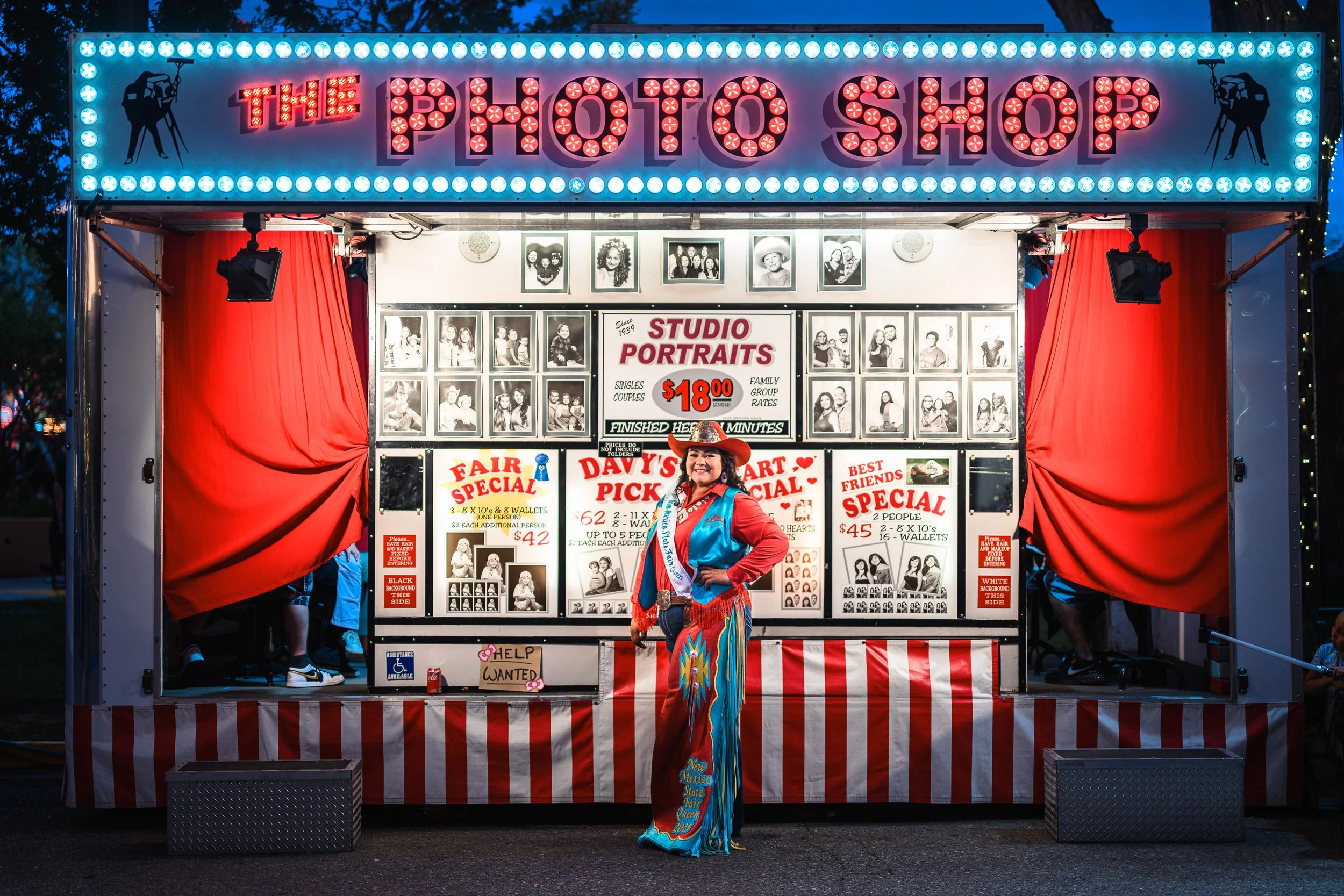 A woman dressed as a rodeo queen standing in front of a night photo stand at a fair, with a sign for the photo shop above. The stand has red and white striped curtains, various photographs on display, and signs advertising portrait packages, includin