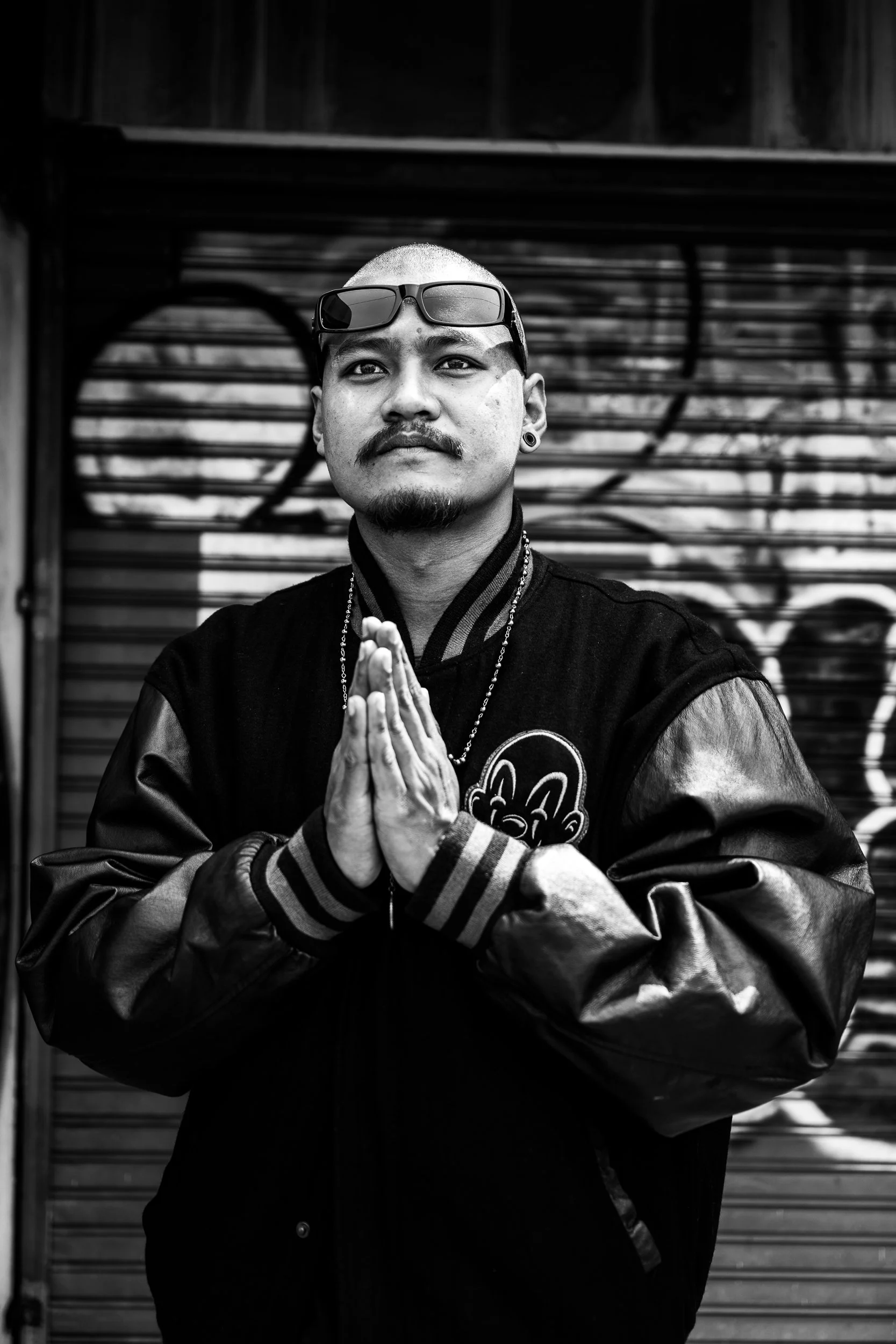 A man with glasses on his head and tattoos on his arm, wearing a varsity jacket, stands with hands pressed together in a prayer or greeting gesture in front of a graffiti-covered metal wall.