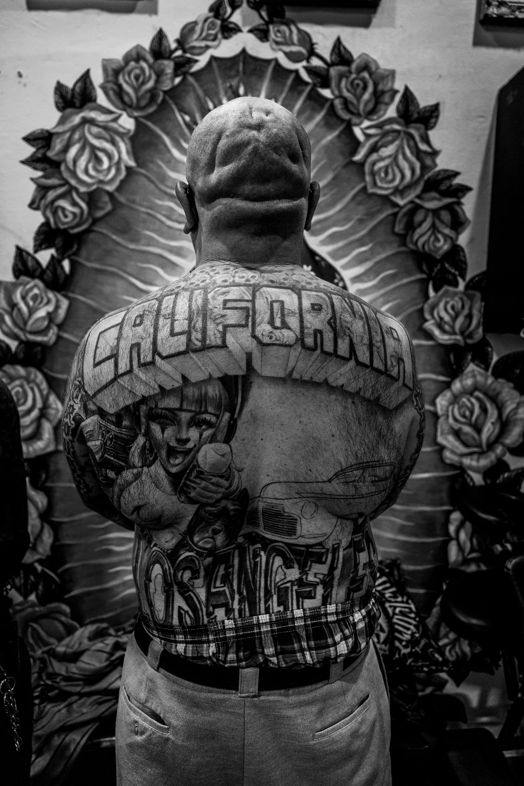 A person with tattoos on their back, including the word "California," standing in front of a decorated mirror with roses.