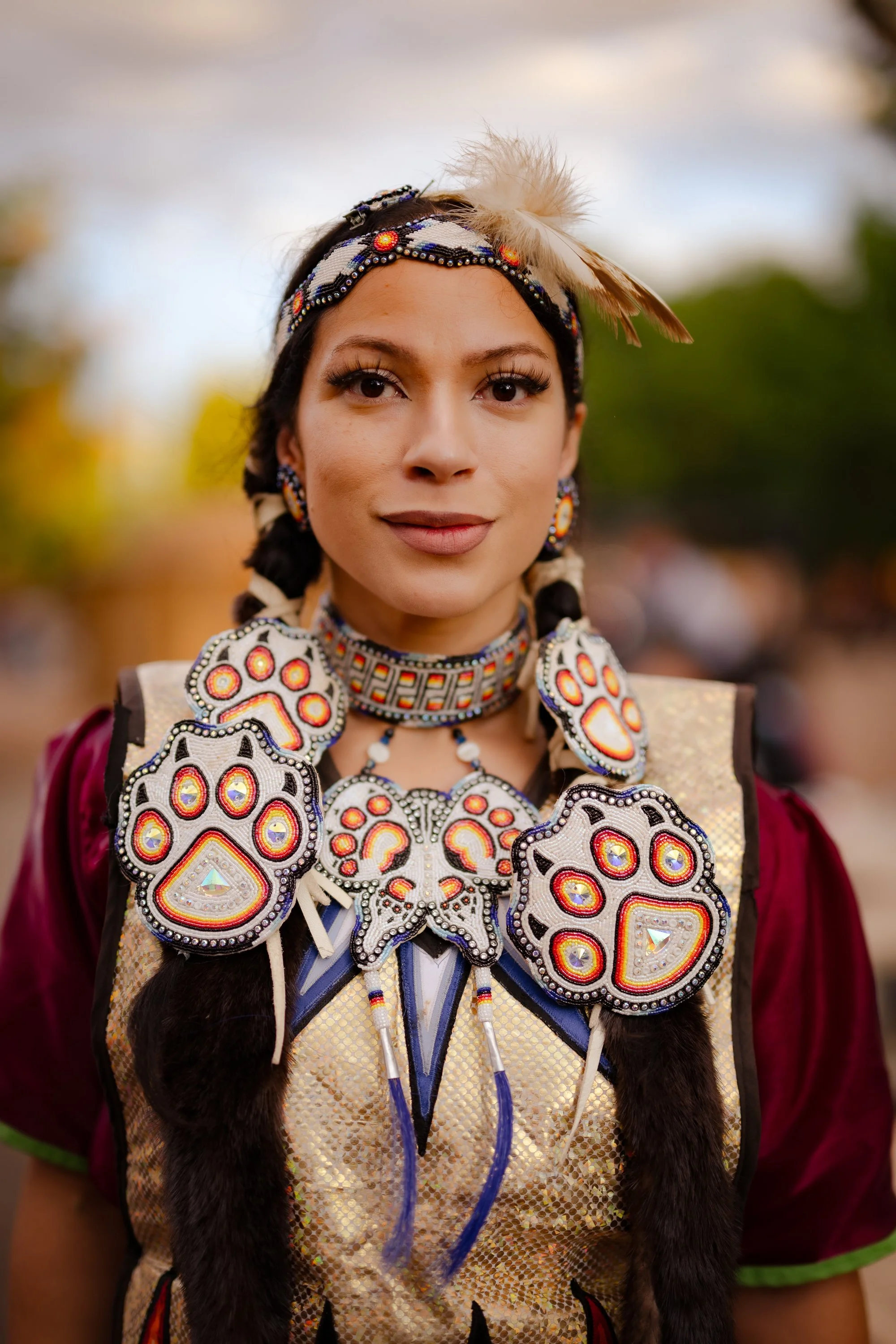 A woman dressed in traditional Native American attire, wearing beaded jewelry with colorful patterns, feathers in her hair, and a decorated vest, standing outdoors with blurred trees and sky in the background.