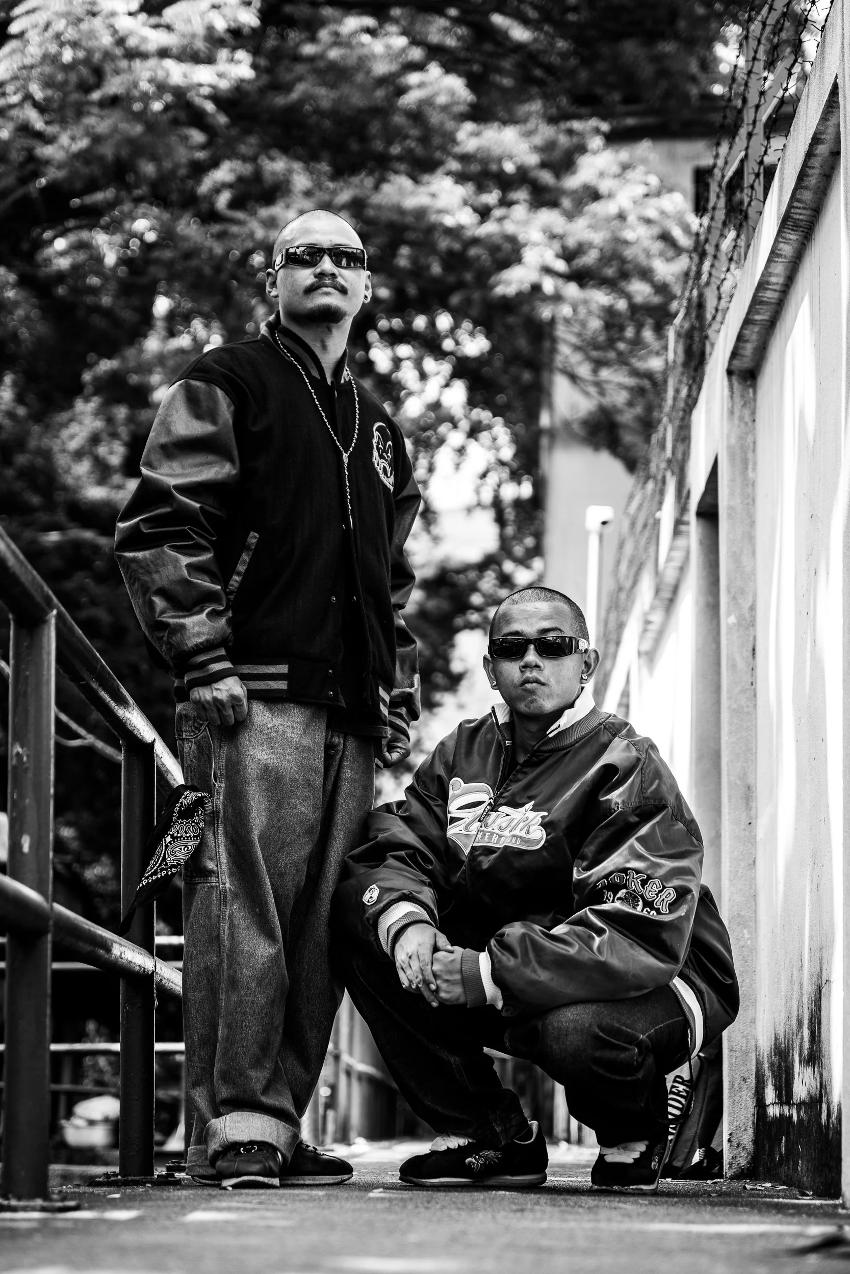 Two men with sunglasses, dressed in streetwear, pose on a city sidewalk with trees and a white wall in the background. One man is standing, the other is squatting.