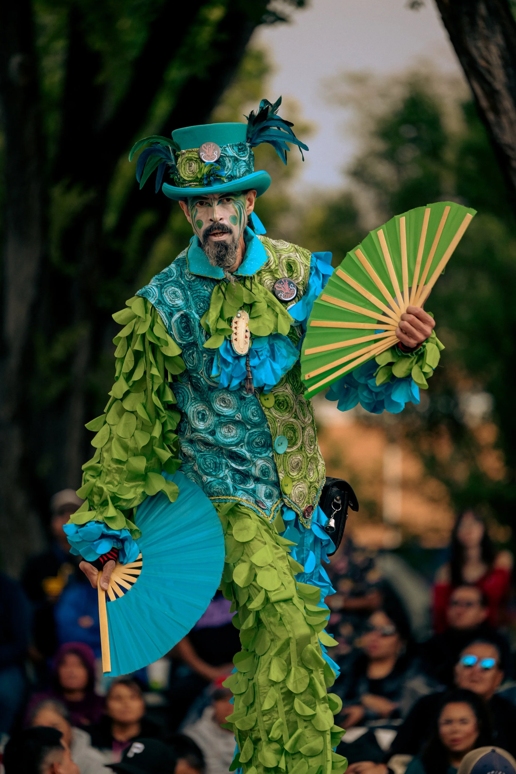 A performer dressed in a colorful, elaborate costume with green and blue fabric, feathers, and accessories, holding a green fan, standing outdoors with a crowd watching.