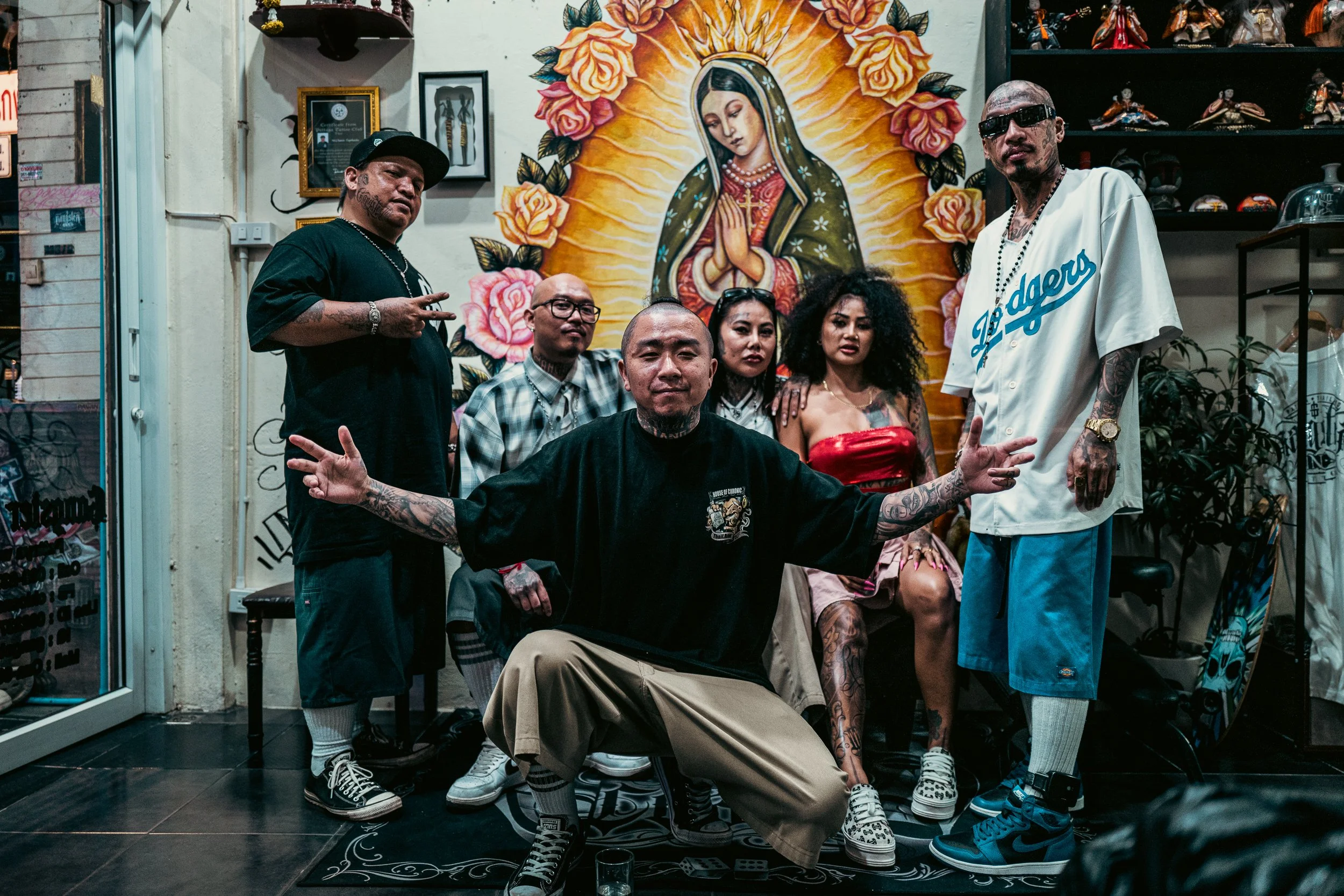 A group of seven diverse people posing in front of a religious icon of the Virgin Mary surrounded by roses, inside a decorated room with shelves of figurines, art, and plants.