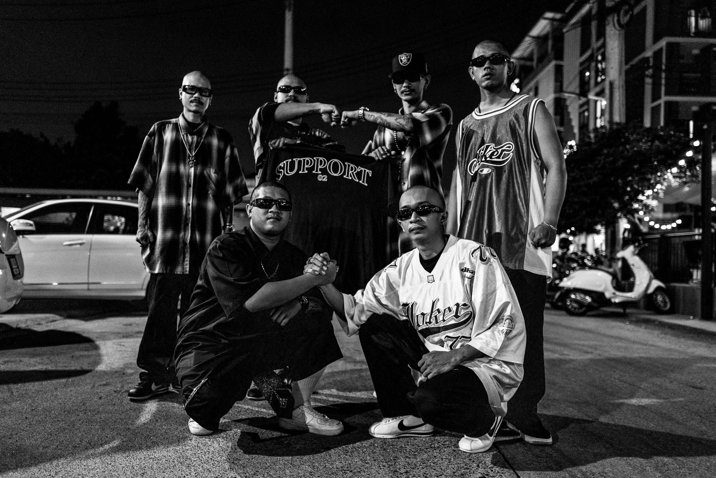 Group of young men posing on a city street at night, some wearing sunglasses and sports jerseys, with motorcycles and cars in the background.