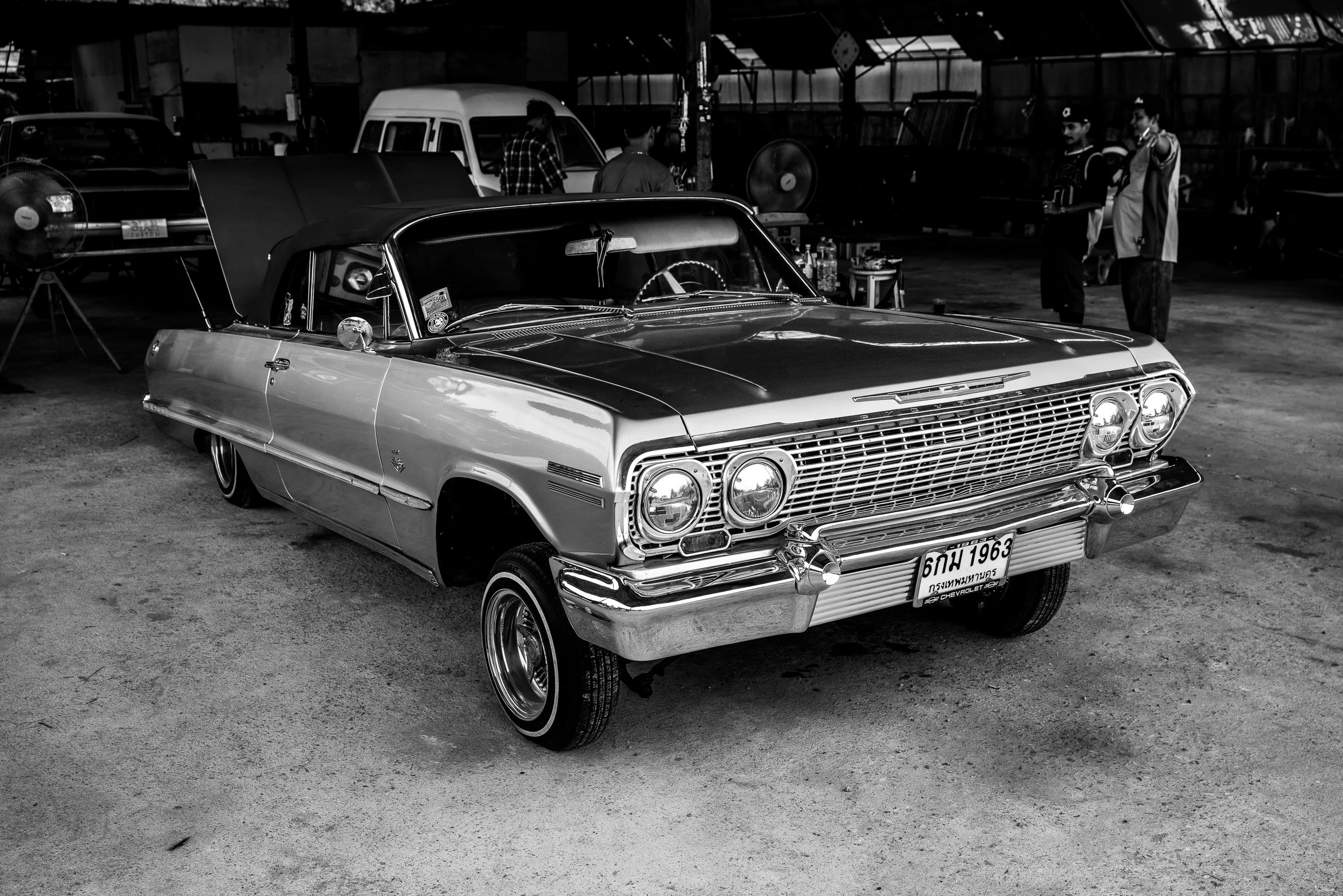 A vintage 1963 Chevrolet car with a black convertible top in a garage with other vehicles and people in the background.
