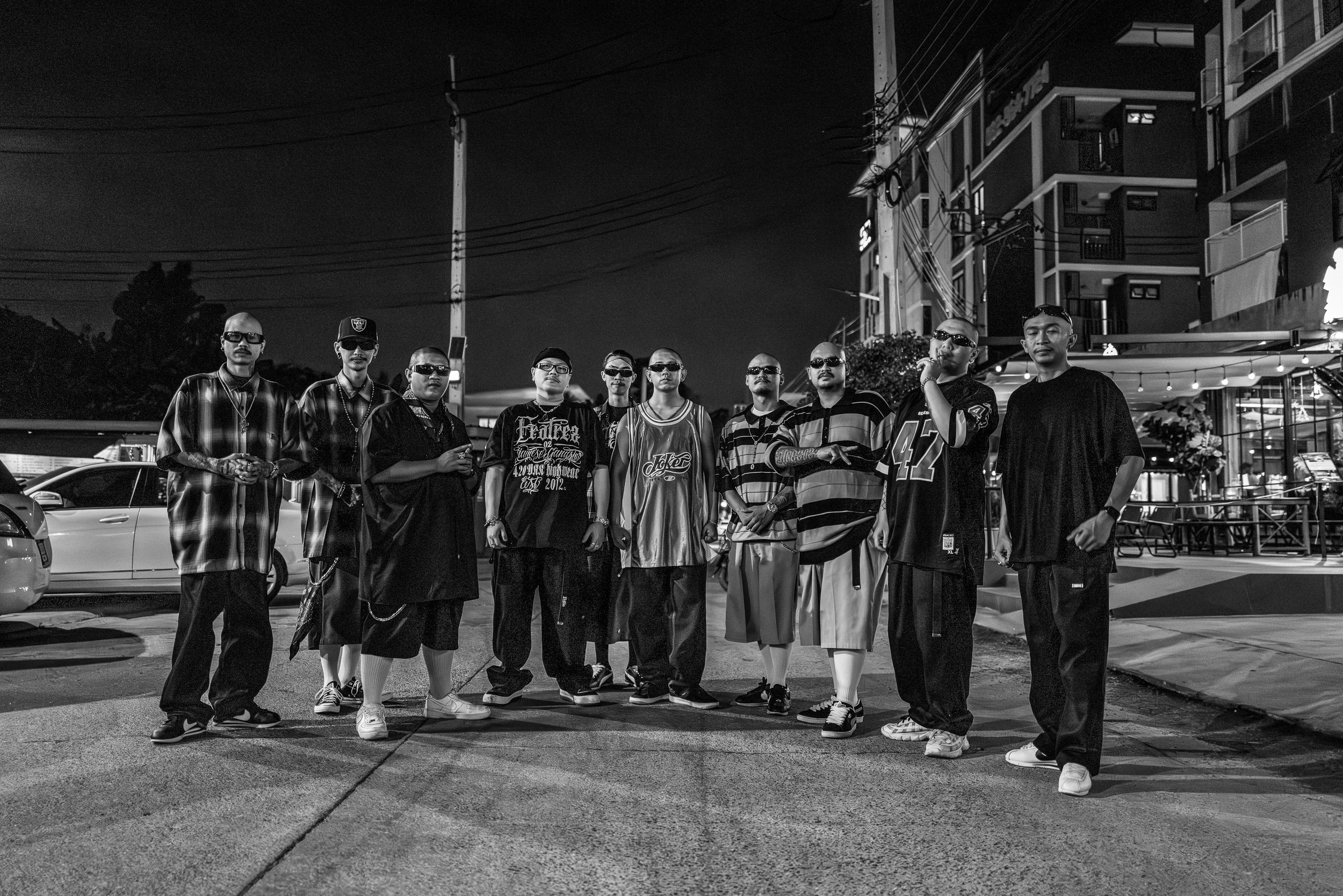 A group of eleven men standing on a street at night, dressed in casual streetwear, with some wearing sunglasses and caps, in an urban neighborhood with buildings and cars in the background.