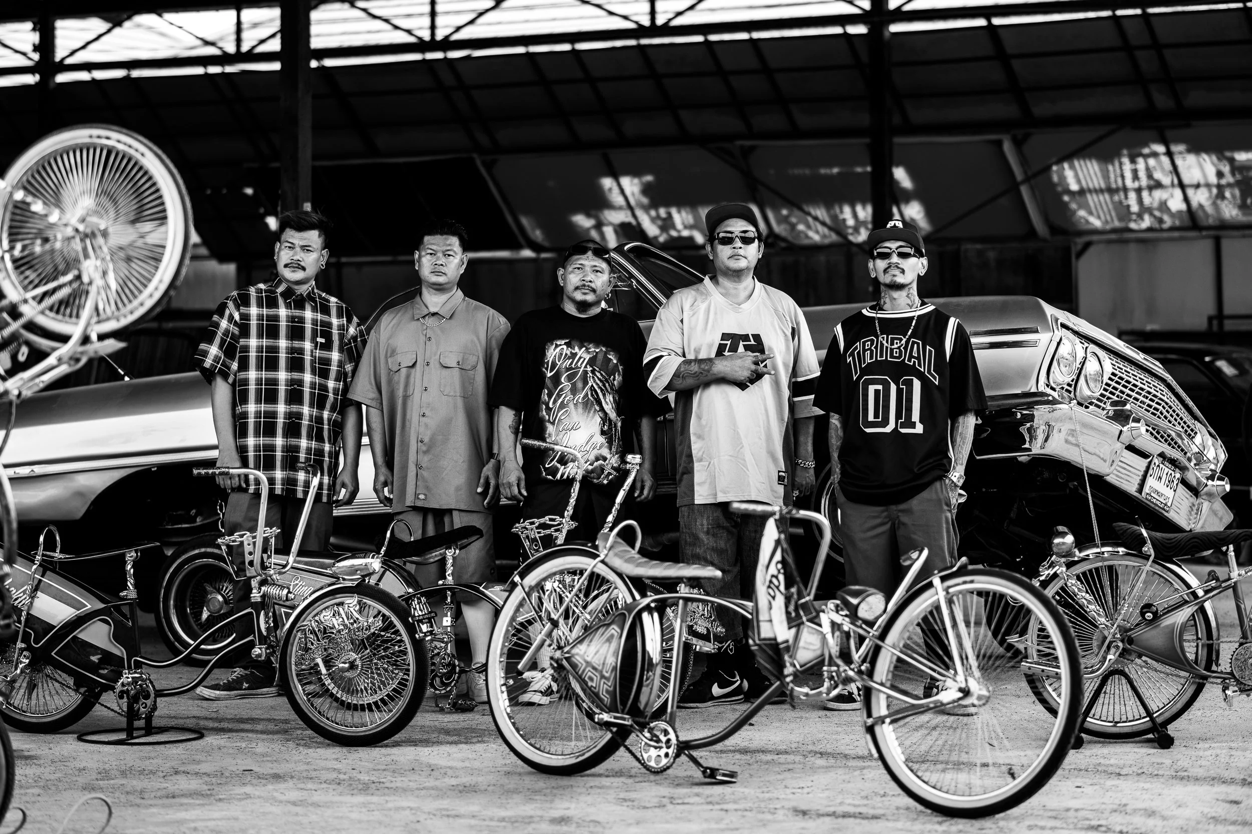 A group of five men standing in a row behind vintage bicycles and classic cars, in a setting with a large roof structure.