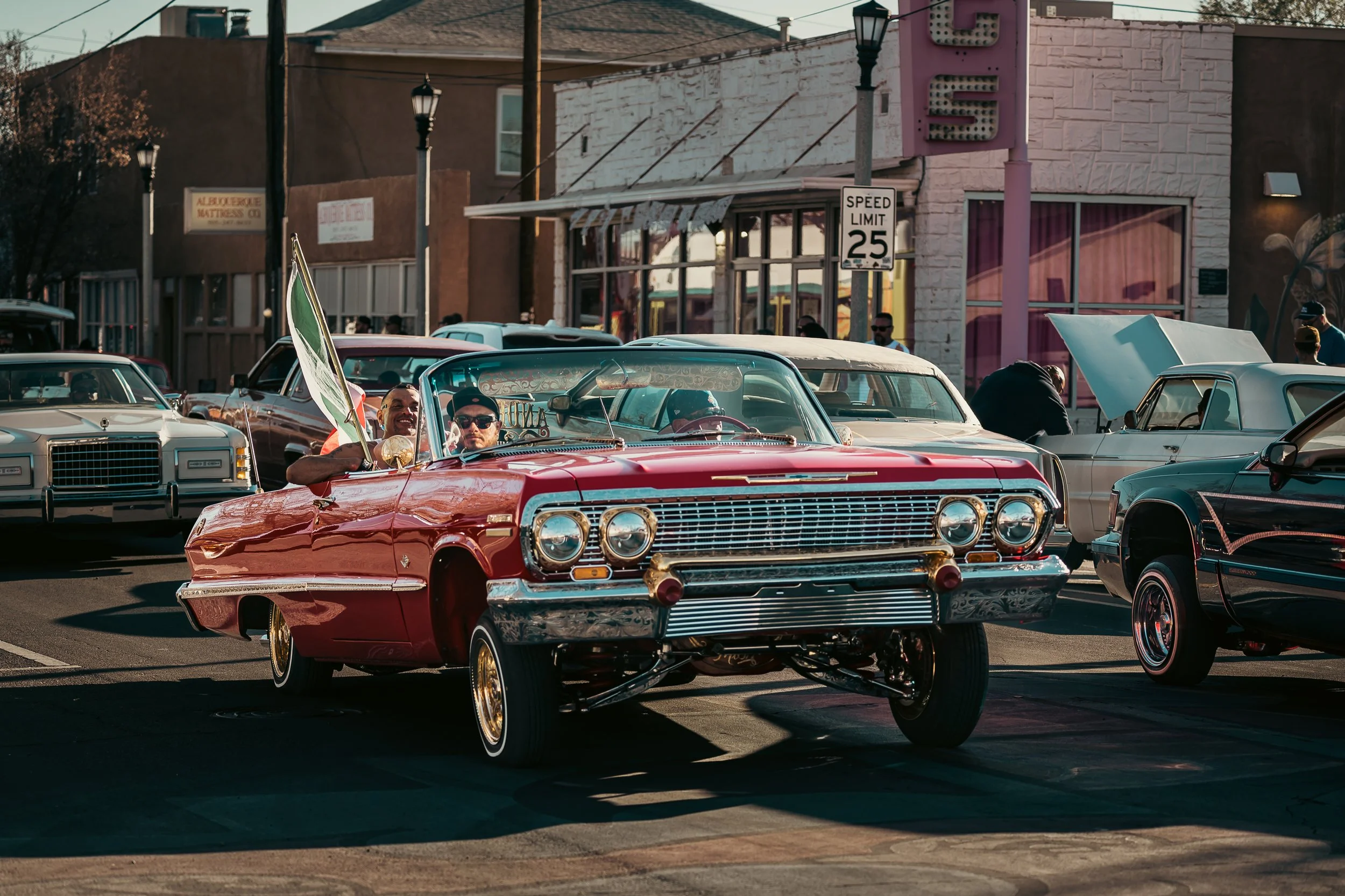 A vintage red Chevrolet convertible car with two men inside, one holding an Italian flag, parked among other classic cars on a city street during a car show or parade.