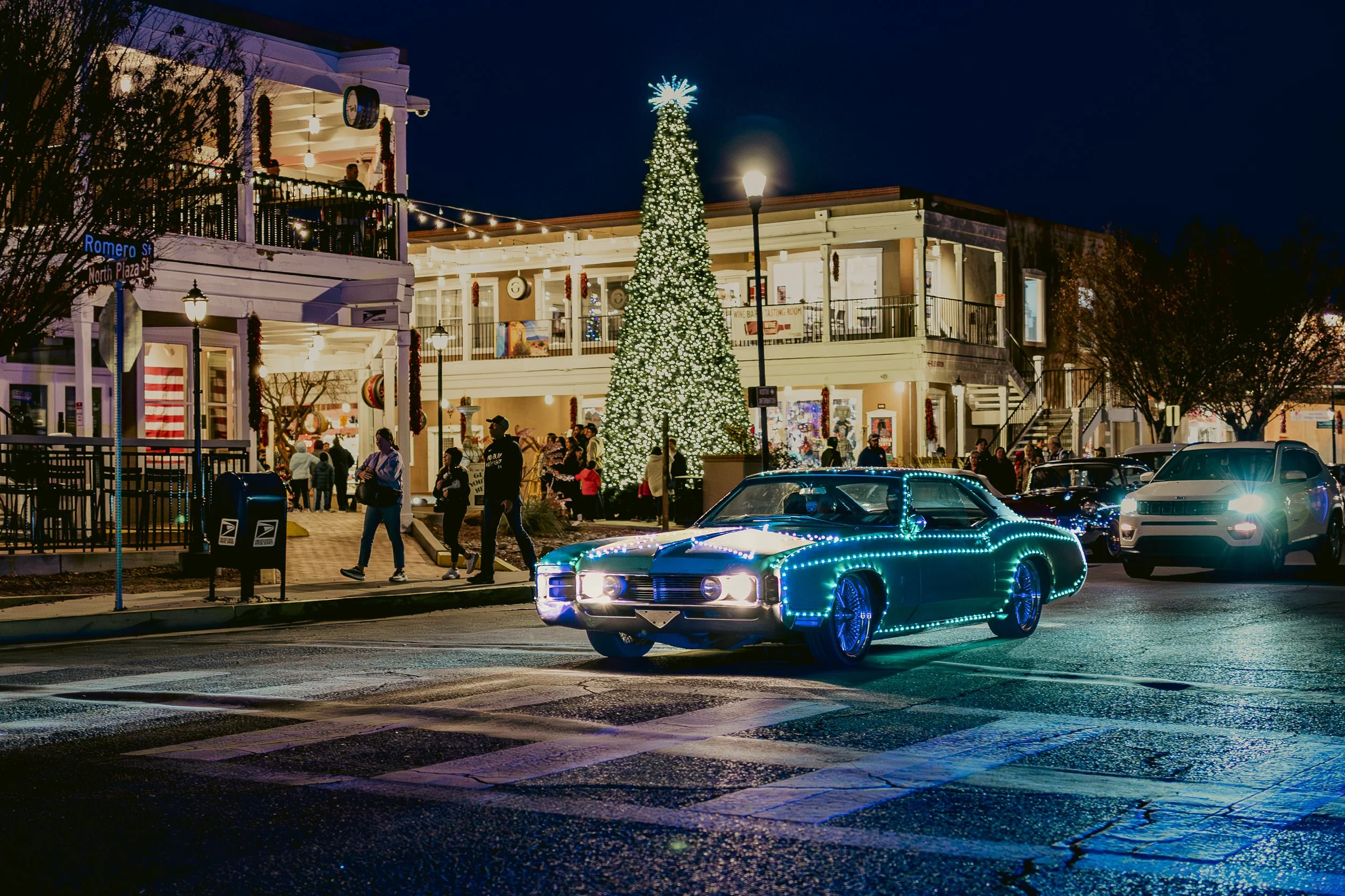 A decorated Christmas tree with white lights in a town square at night. Classic cars with blue LED lights are parked on the street. People are walking and socializing outside storefronts decorated for the holidays.