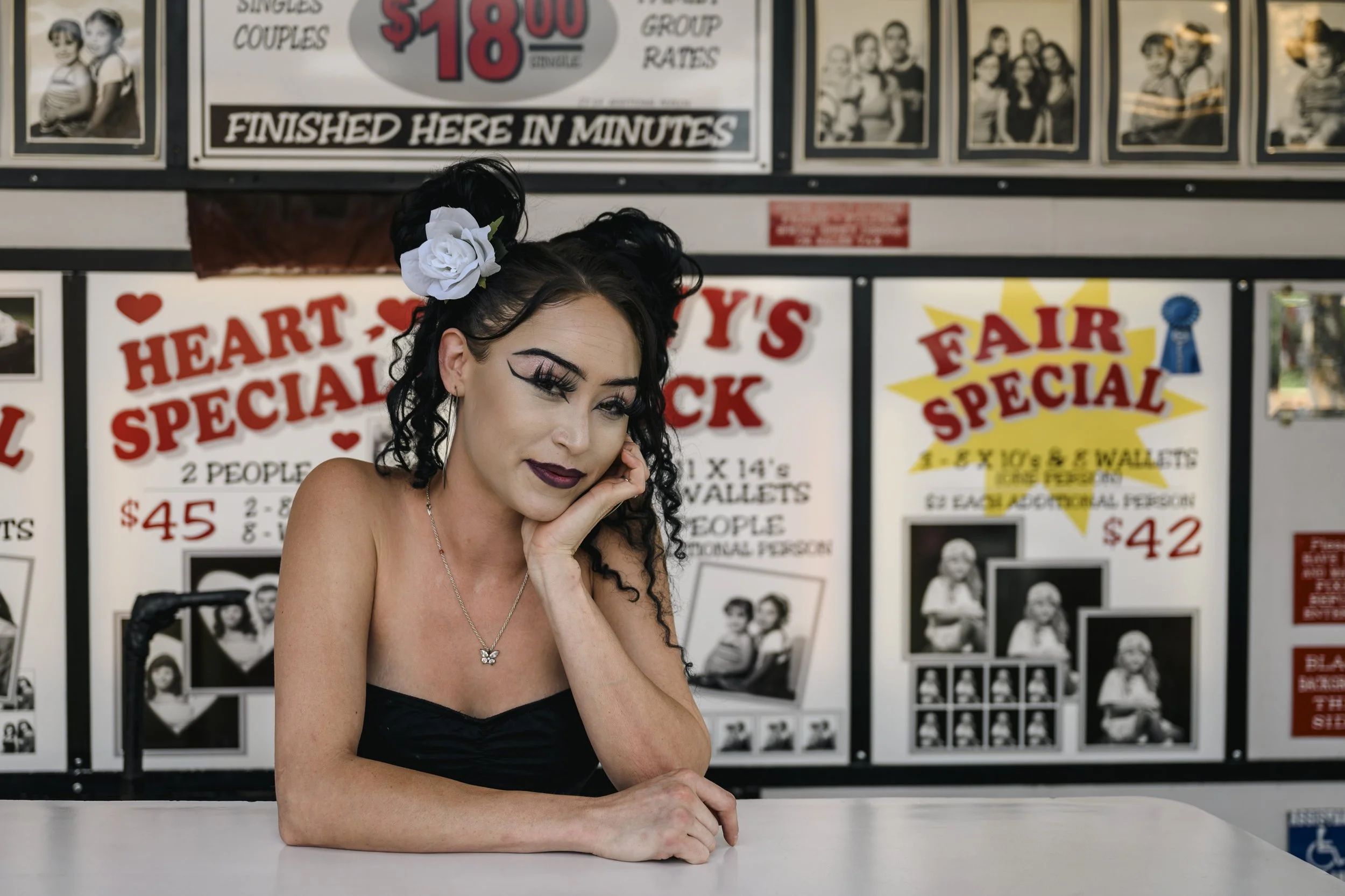 A woman with dark, curly hair styled in two buns decorated with white roses, wearing dark makeup and a black top, sitting at a table with her head resting on her hand, in front of a background with various colorful poster signs advertisement for a fair or carnival.