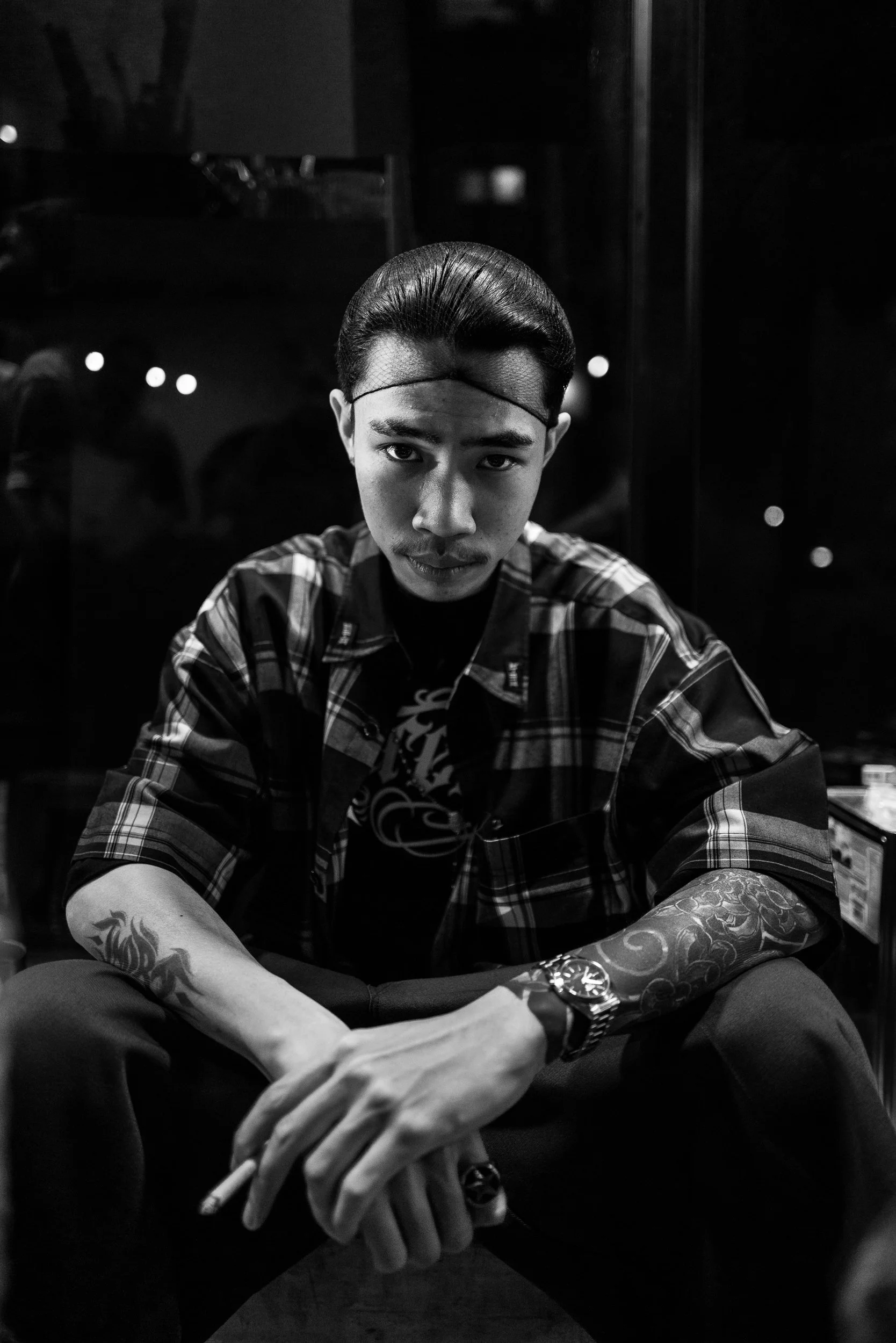A young man with tattoos on his arms, wearing a plaid shirt and a headband, sitting and looking into the camera in a dimly lit room.