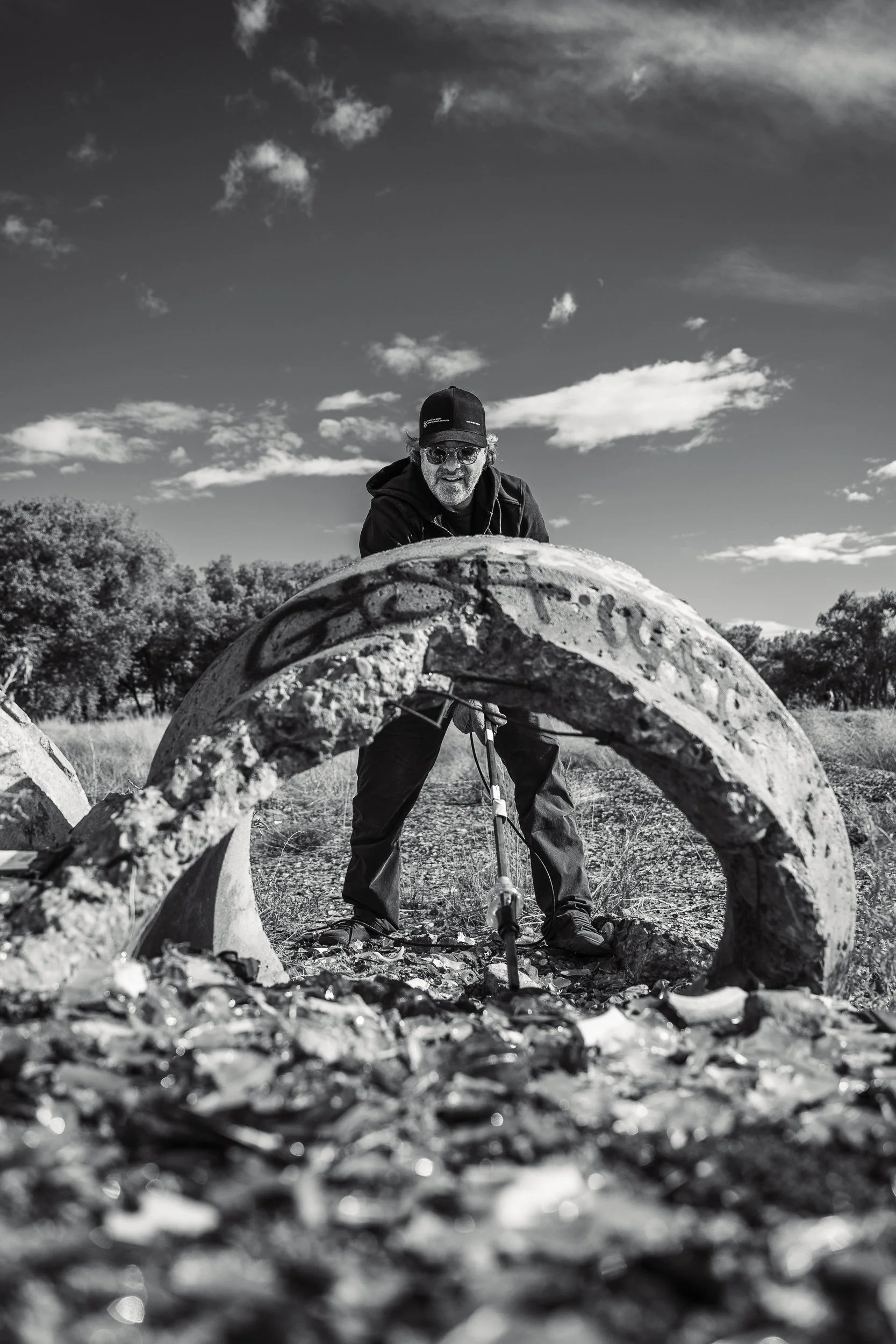 A man in sunglasses and a cap using a metal detector outdoors, with large, curved concrete structures and scattered debris on the ground under a partly cloudy sky.