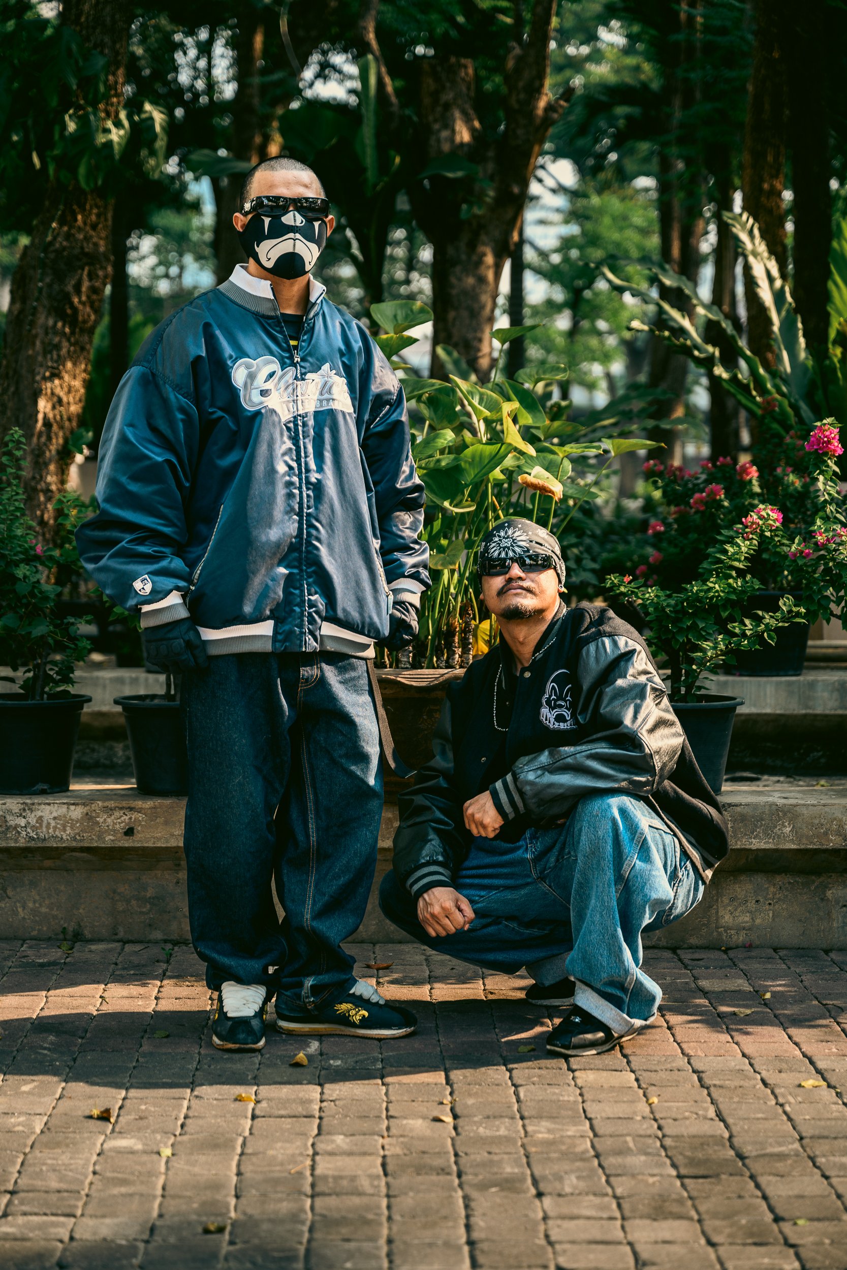 Two men in urban streetwear pose outdoors with potted plants and trees in the background. One man is standing and the other is squatting. Both wear jackets, sunglasses, and bandanas.