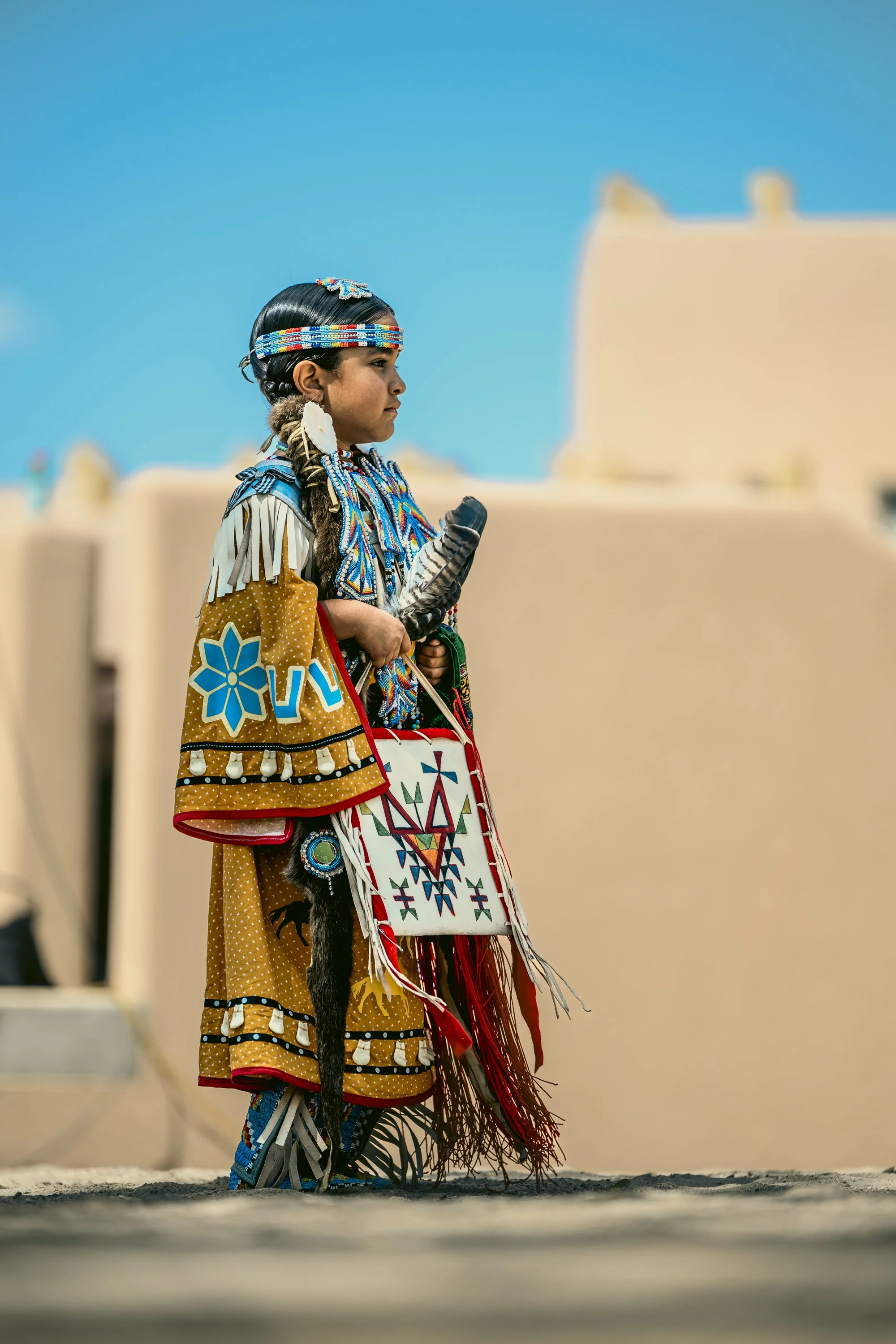 Young Native American girl in traditional clothing holding a beaded object, standing outdoors with adobe buildings and a clear blue sky in the background.