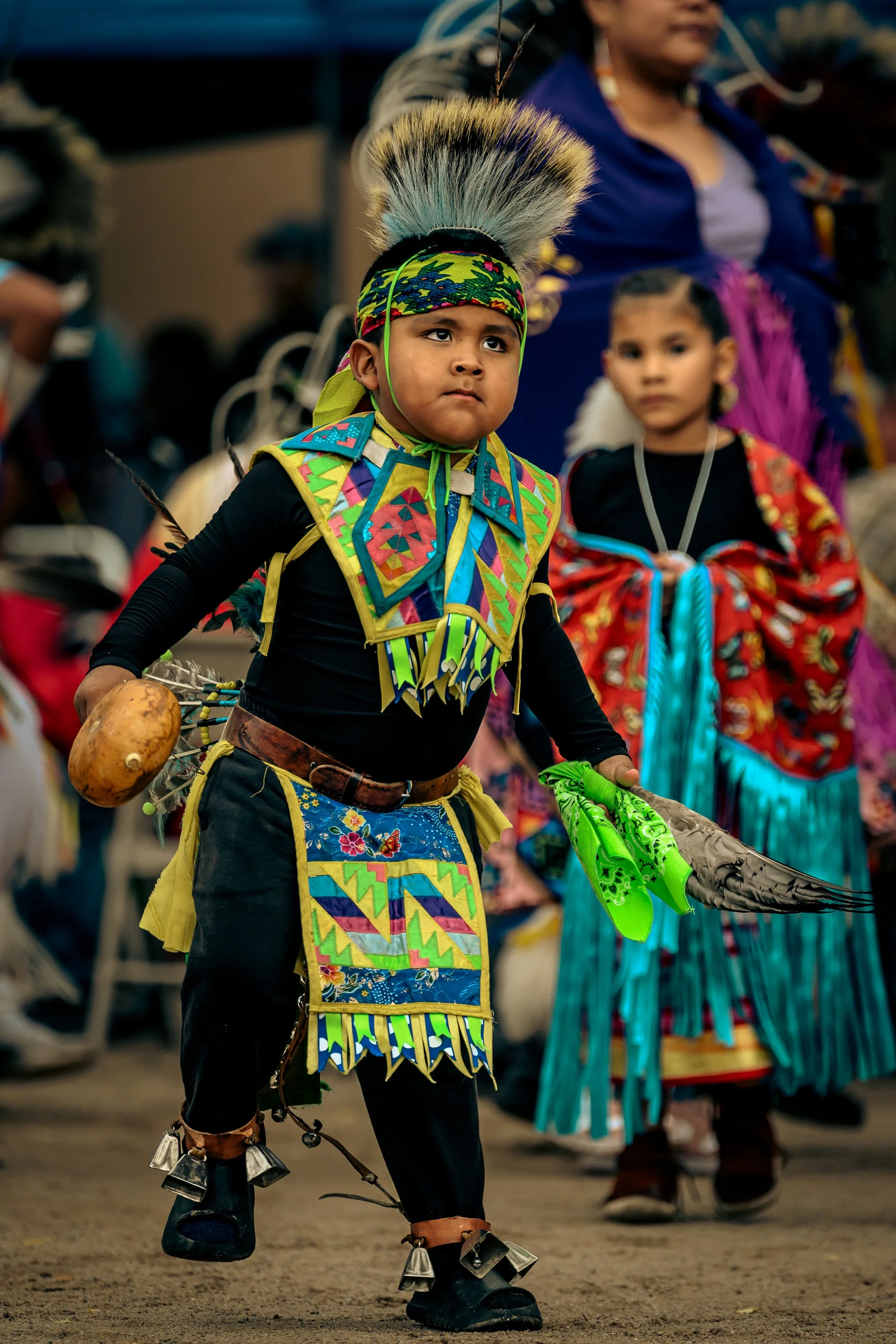 A young boy dressed in traditional Native American attire, including a feathered headdress, colorful beaded clothing, and bells on his shoes, participating in a cultural dance or celebration.
