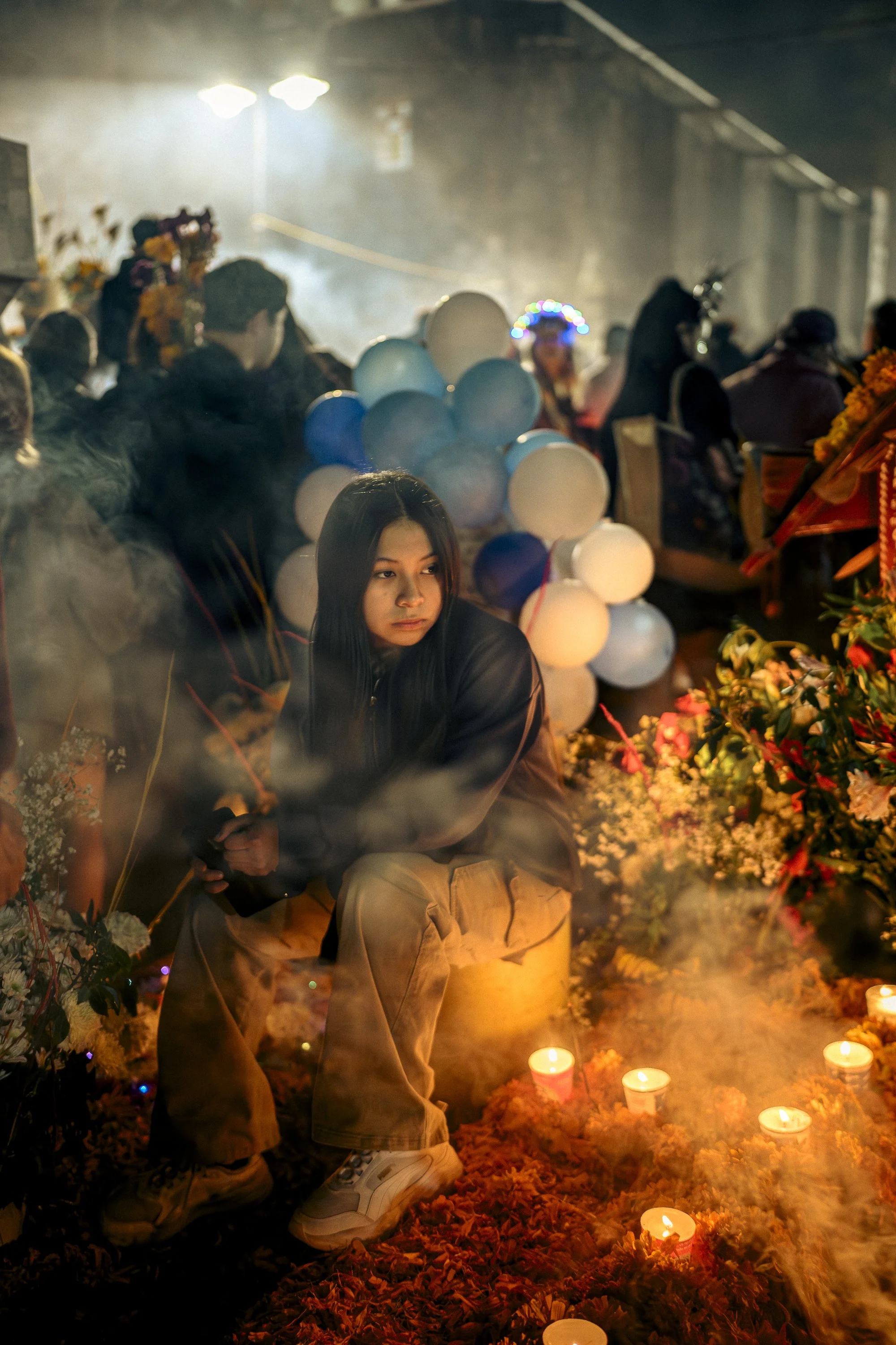 A woman sitting among flowers, candles, and balloons during a nighttime outdoor event, with smoke and dim lighting.