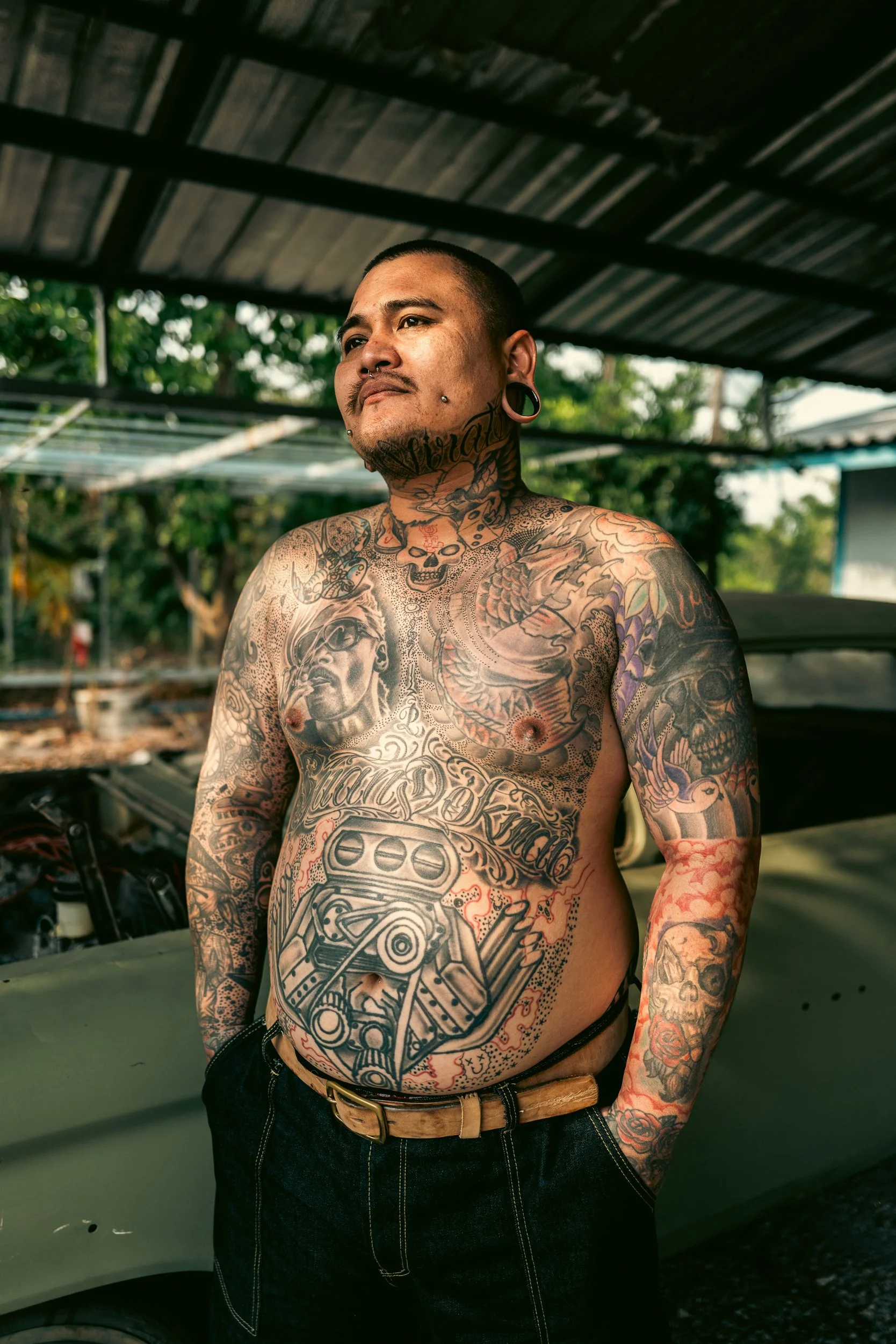 A tattooed man with tattoos covering his chest, arms, and neck, standing outdoors under a covered structure, with a vehicle and green trees in the background.