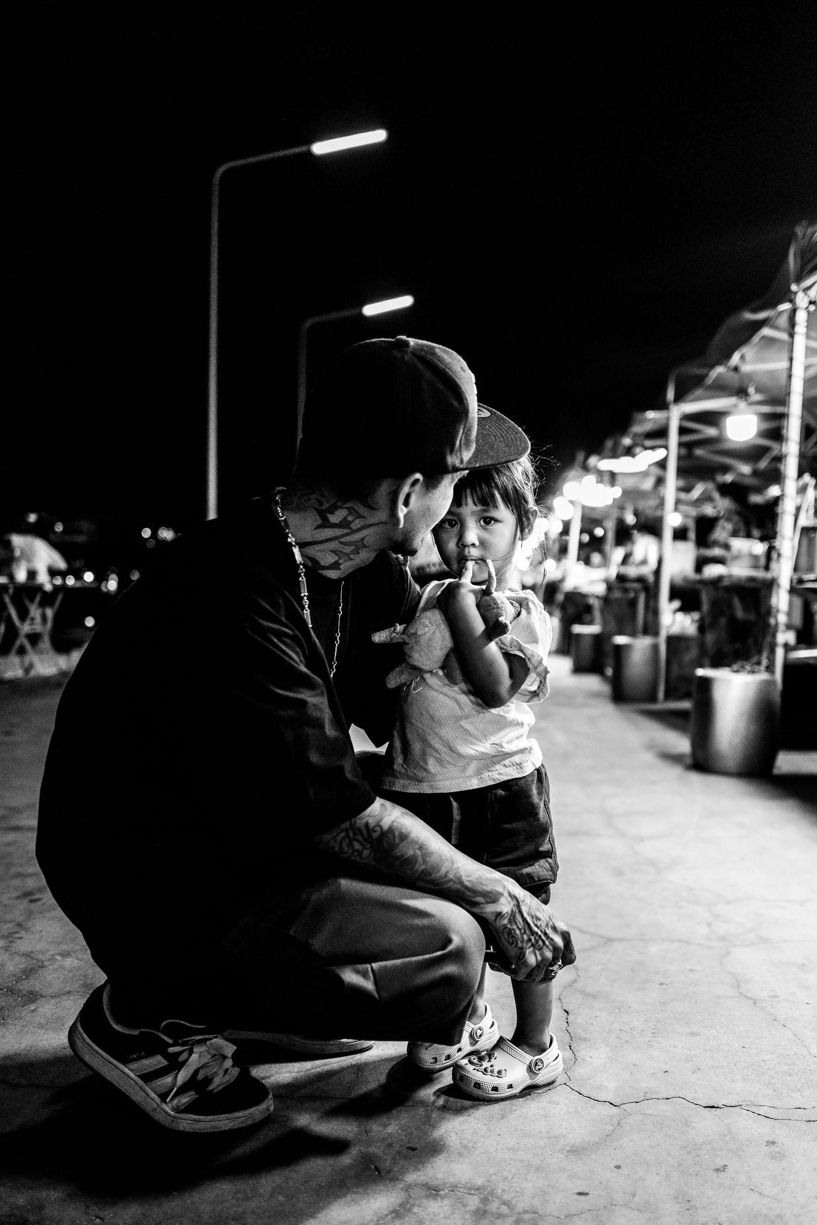 A man with tattoos, wearing a cap and chain, kneeling and holding a young girl with a pacifier at a night market. The girl has short hair, wearing a dress and Crocs, looking at the camera. Market stalls with lights line the background.
