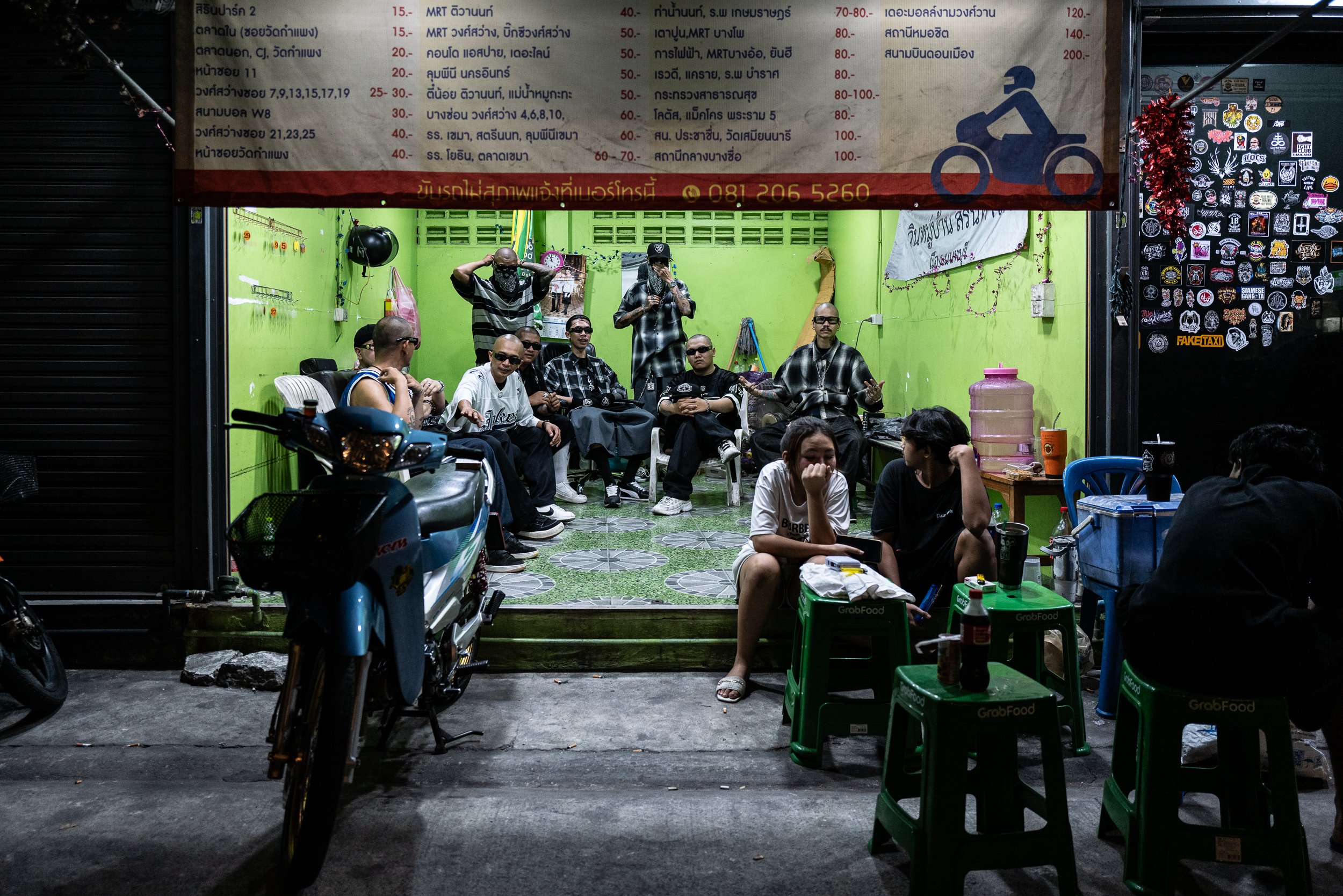 A group of men wearing sunglasses and casual clothes sitting inside a small brightly painted green room, some standing and some seated, with one woman and a man sitting outside at small tables with food and drinks, and a motorcycle parked in front.
