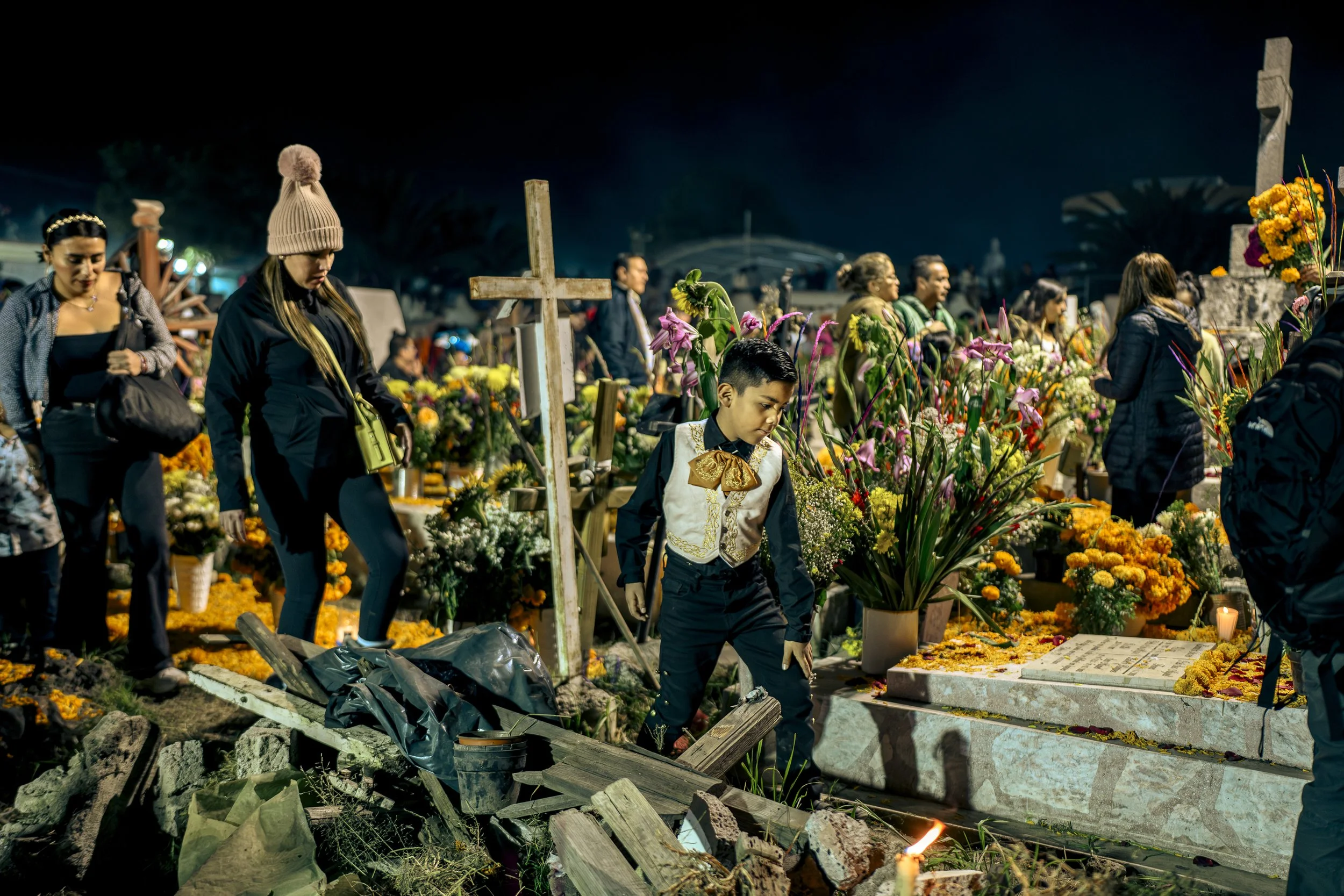 People paying respects at a nighttime cemetery with flowers and candles, some looking solemn, others reflecting.