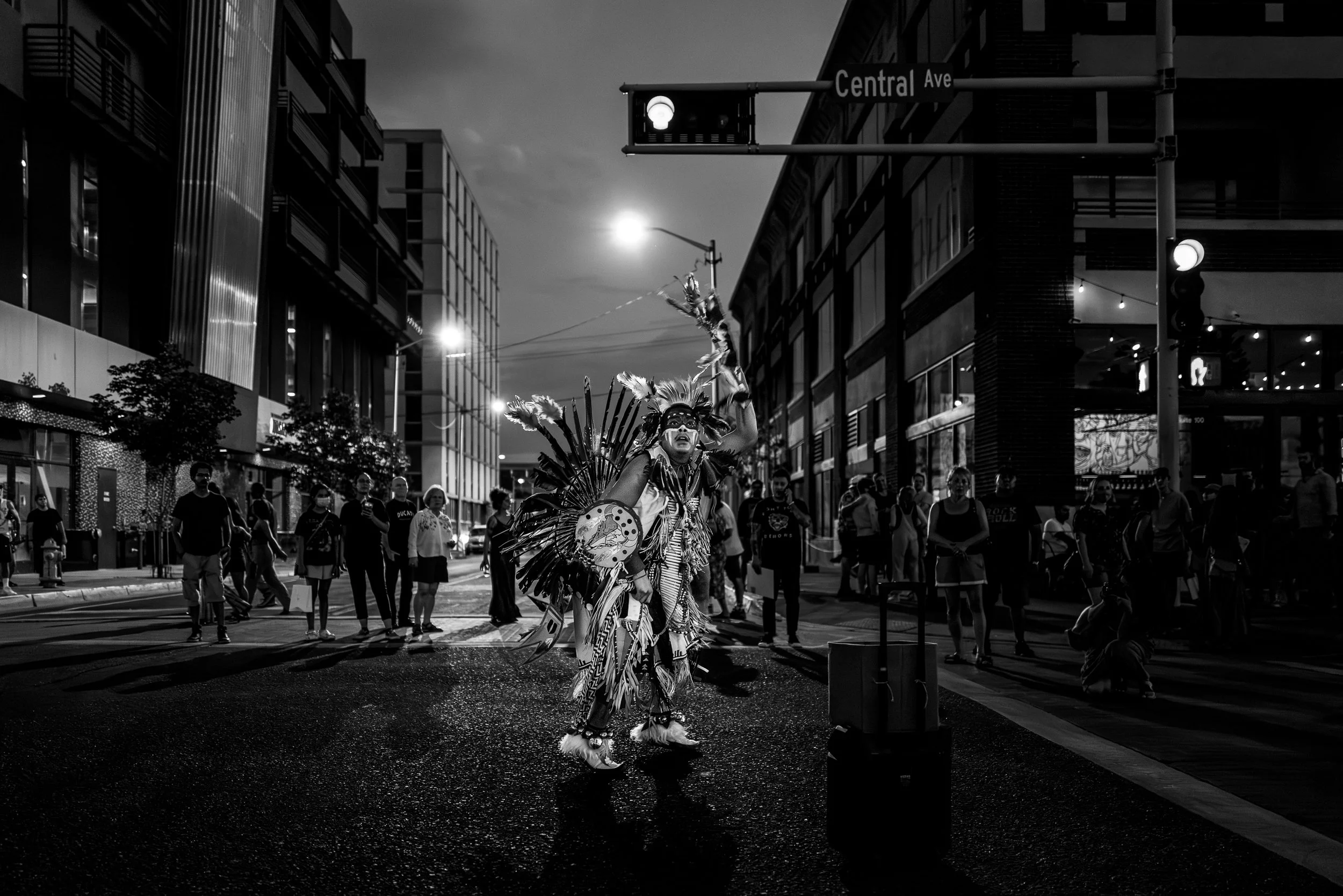Black-and-white photograph of a dancer in regalia crossing Central Avenue during a post-COVID gathering in downtown Albuquerque.