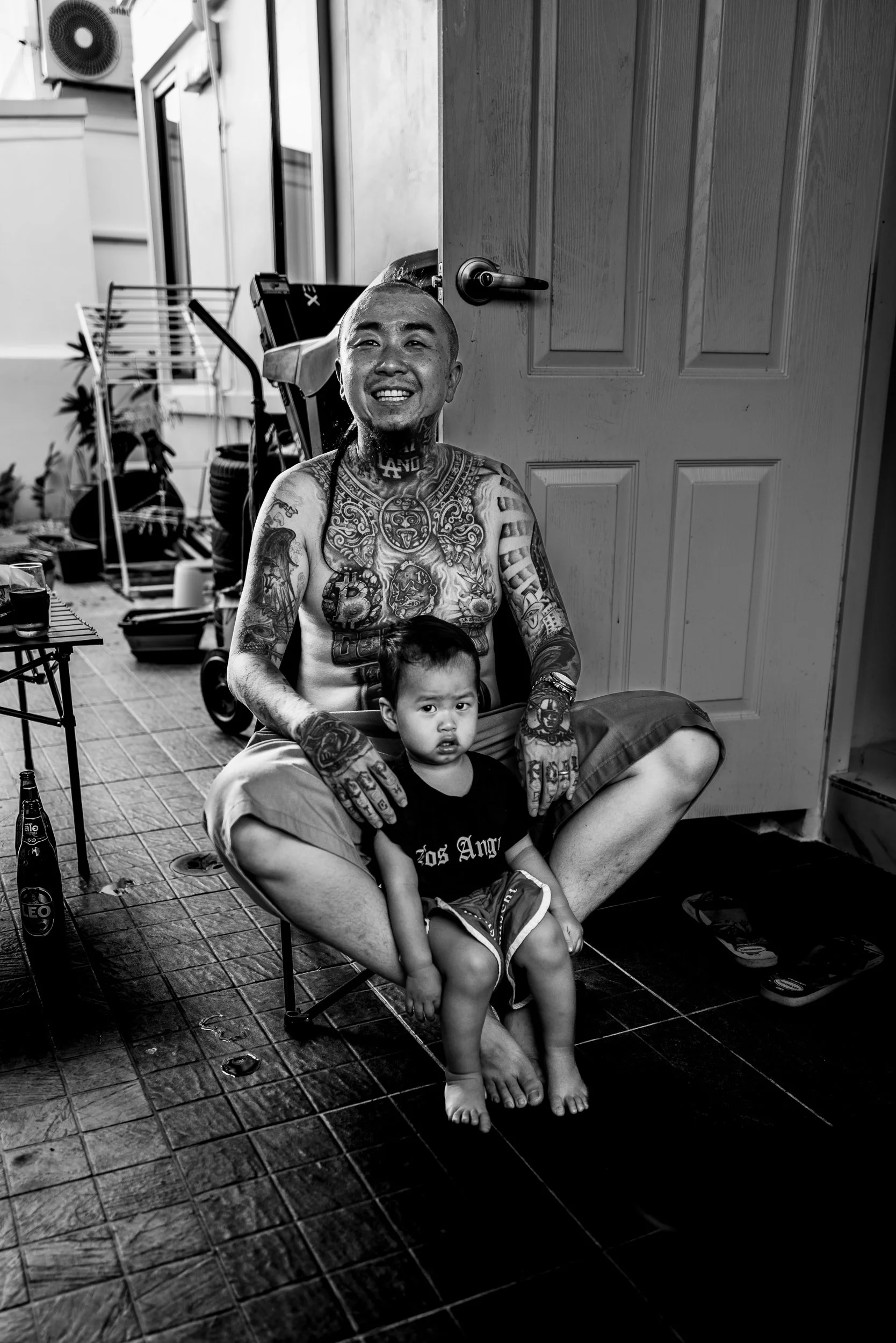 A smiling tattooed man sitting cross-legged on the floor, holding a young child on his lap, both inside a room with a door open to the outside. The man has tattoos on his chest, arms, and neck, and is barefoot. The child has a serious expression and 