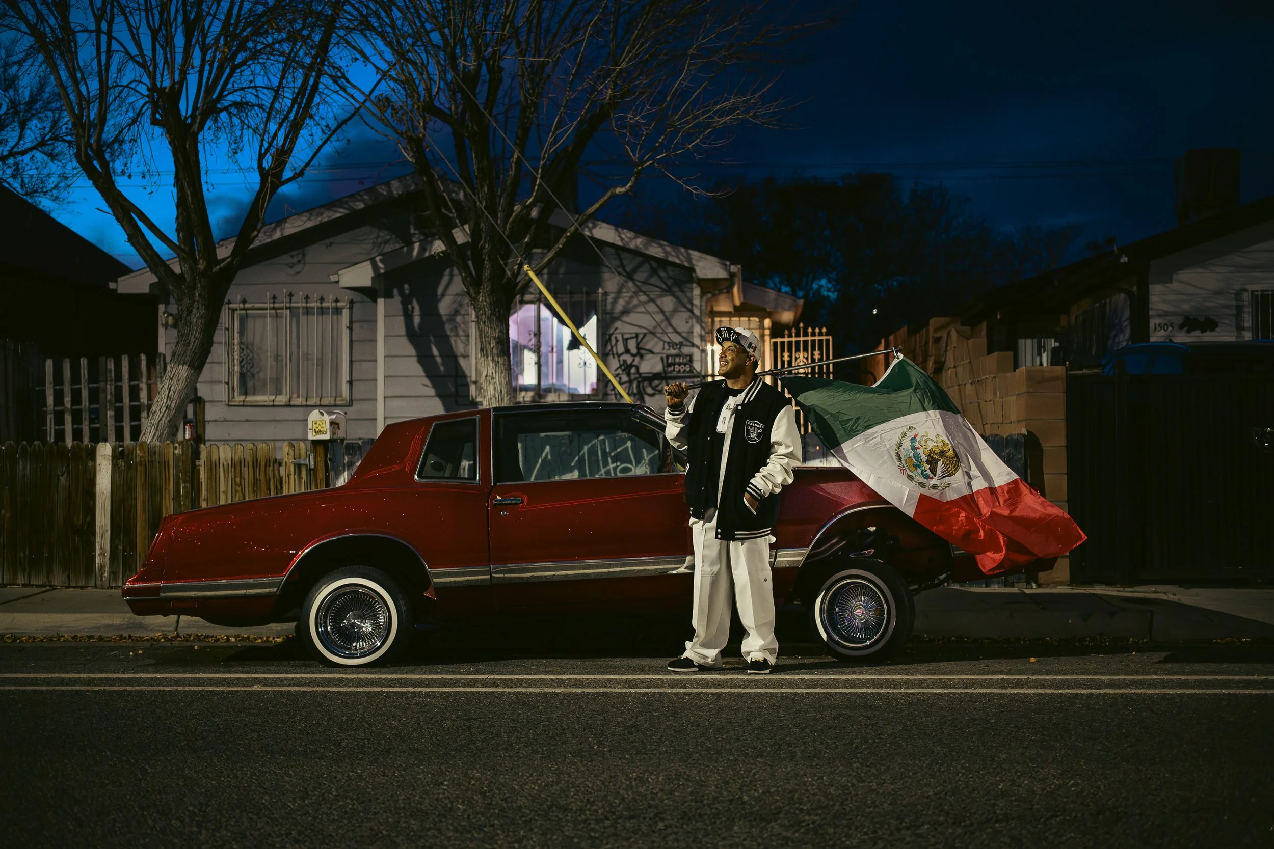 A man standing on the street in front of a red classic car with a Mexican flag attached, under a dark evening sky, with a house and trees in the background.