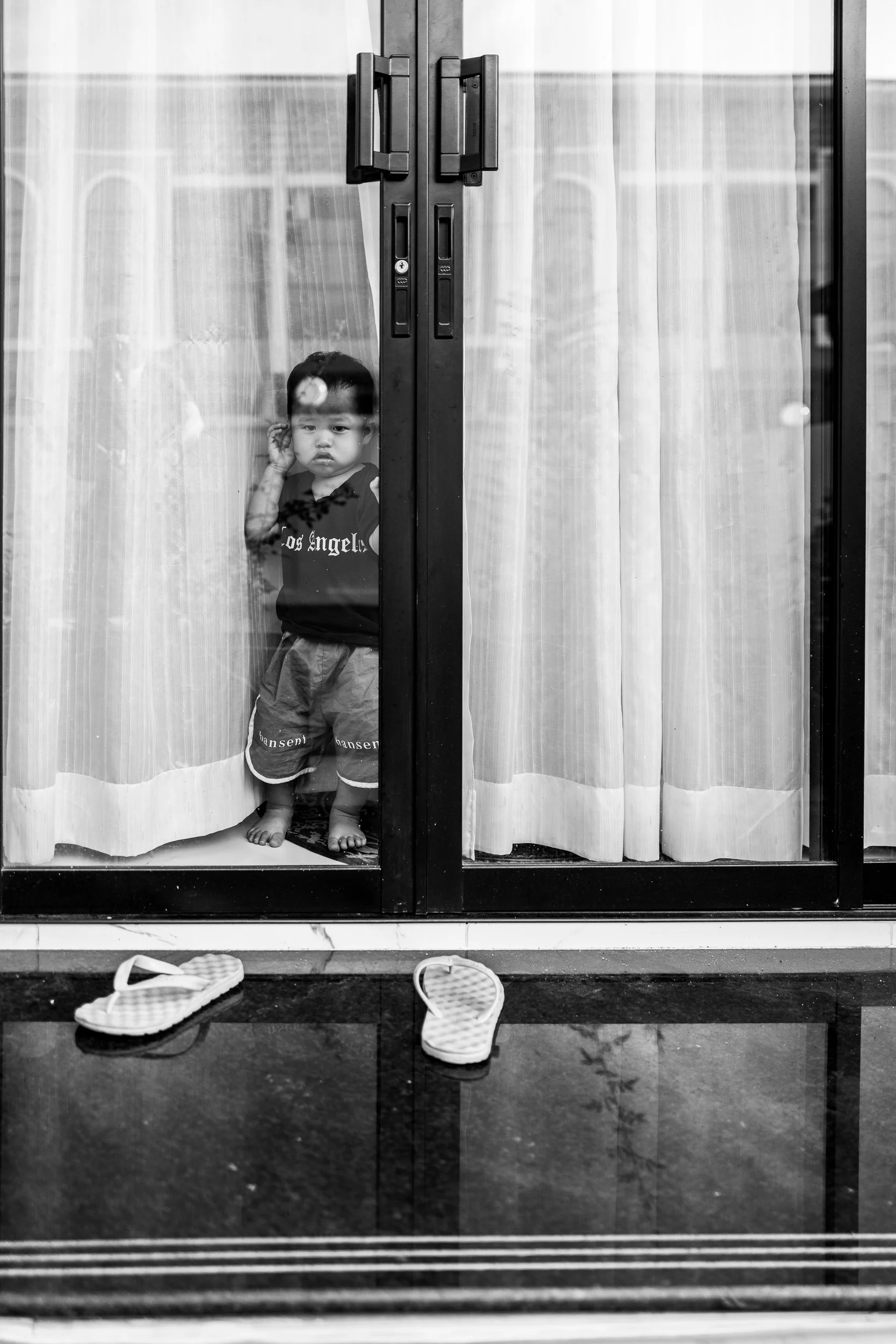A young child standing behind a glass door, looking out. The child is barefoot and wearing a dark t-shirt and shorts. Two slippers are placed on the floor outside the door.